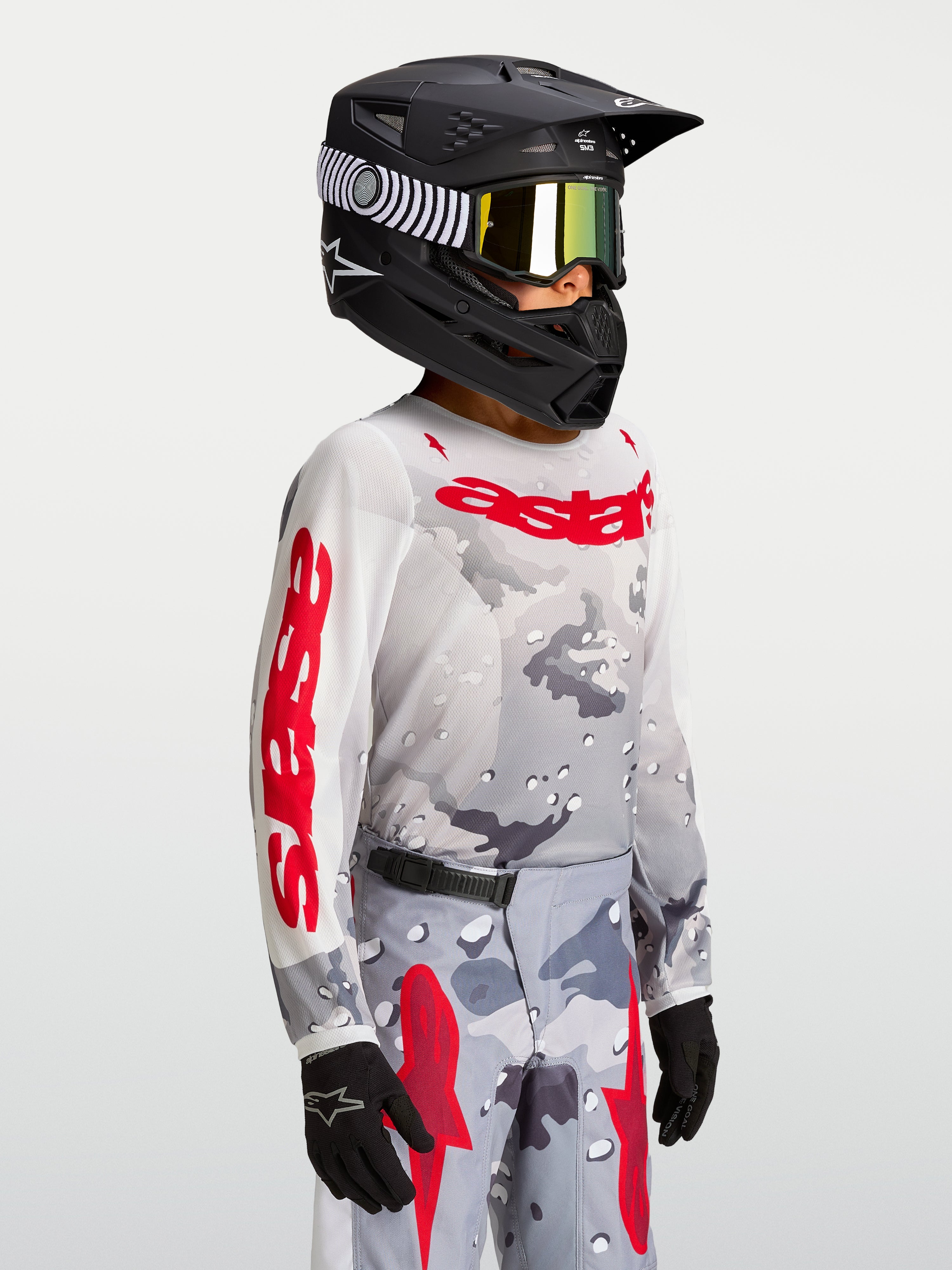 Alpinestars Camisola Fluid Lone Jovem 2027, camisola de motocross cinzenta camuflada e vermelha, modelo com capacete.