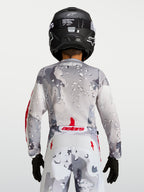 Alpinestars Camisola Fluid Lone Jovem 2027, camisola de motocross para jovem, cinzenta camuflada com detalhes vermelhos, vista traseira com capacete preto.