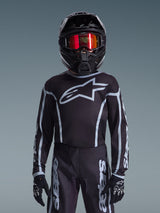 Jovem piloto de motocross com a camisola Alpinestars Youth Fluid Apex 2026 em preto e cinzento, calças a condizer, capacete preto com óculos refletores vermelhos e luvas pretas, equipamento de corrida todo-o-terreno coordenado
