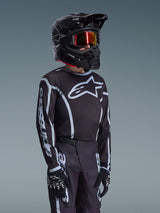 Camisola Alpinestars Youth Fluid Apex 2026 em preto e cinzento, usada por um jovem piloto com um capacete preto a condizer e óculos escurecidos, com a marca Alpinestars em destaque no peito e nas mangas, parte de um conjunto de equipamento de motocross coordenado.