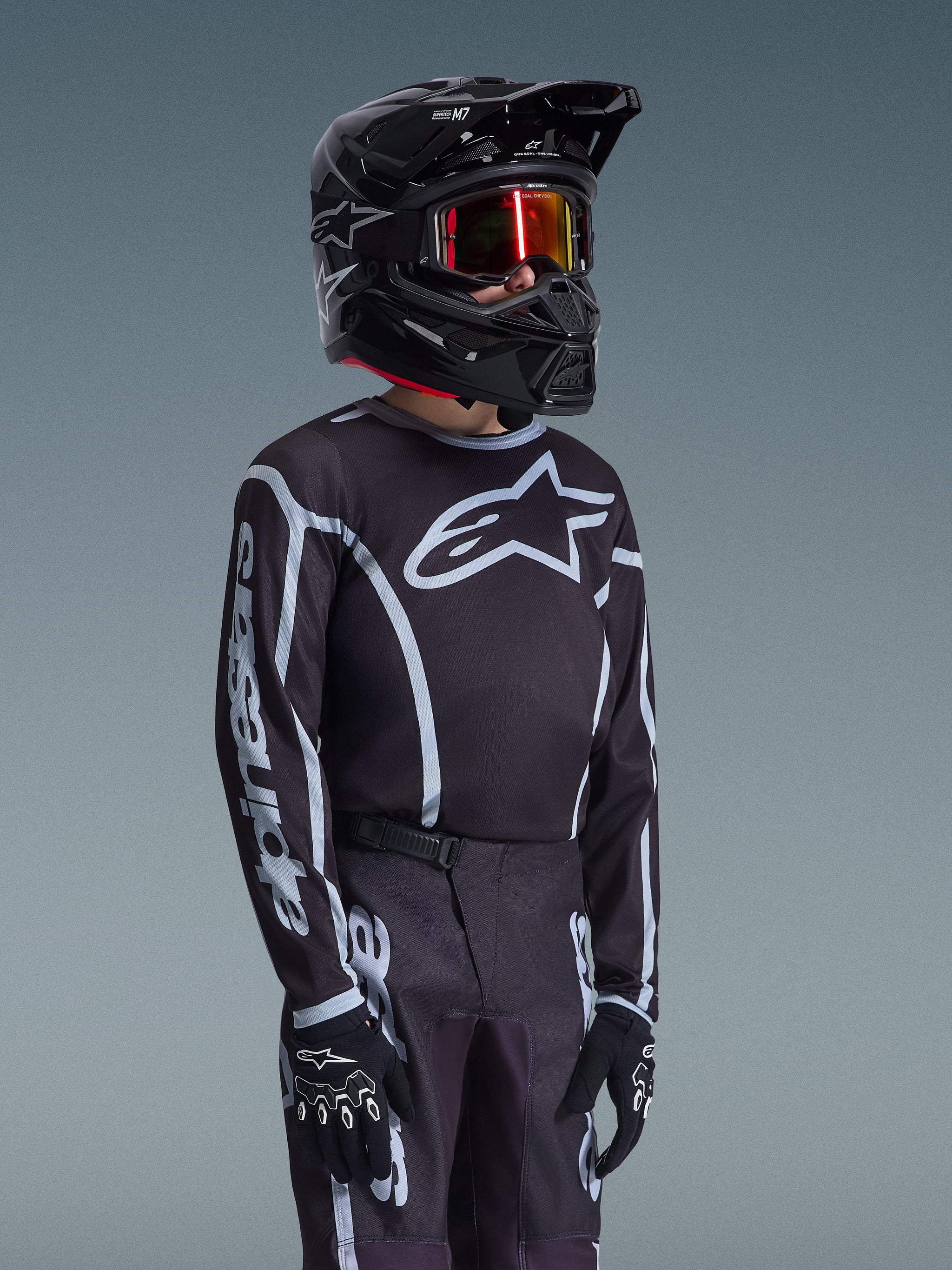 Camisola Alpinestars Youth Fluid Apex 2026 em preto e cinzento, usada por um jovem piloto com um capacete preto a condizer e óculos escurecidos, com a marca Alpinestars em destaque no peito e nas mangas, parte de um conjunto de equipamento de motocross coordenado.