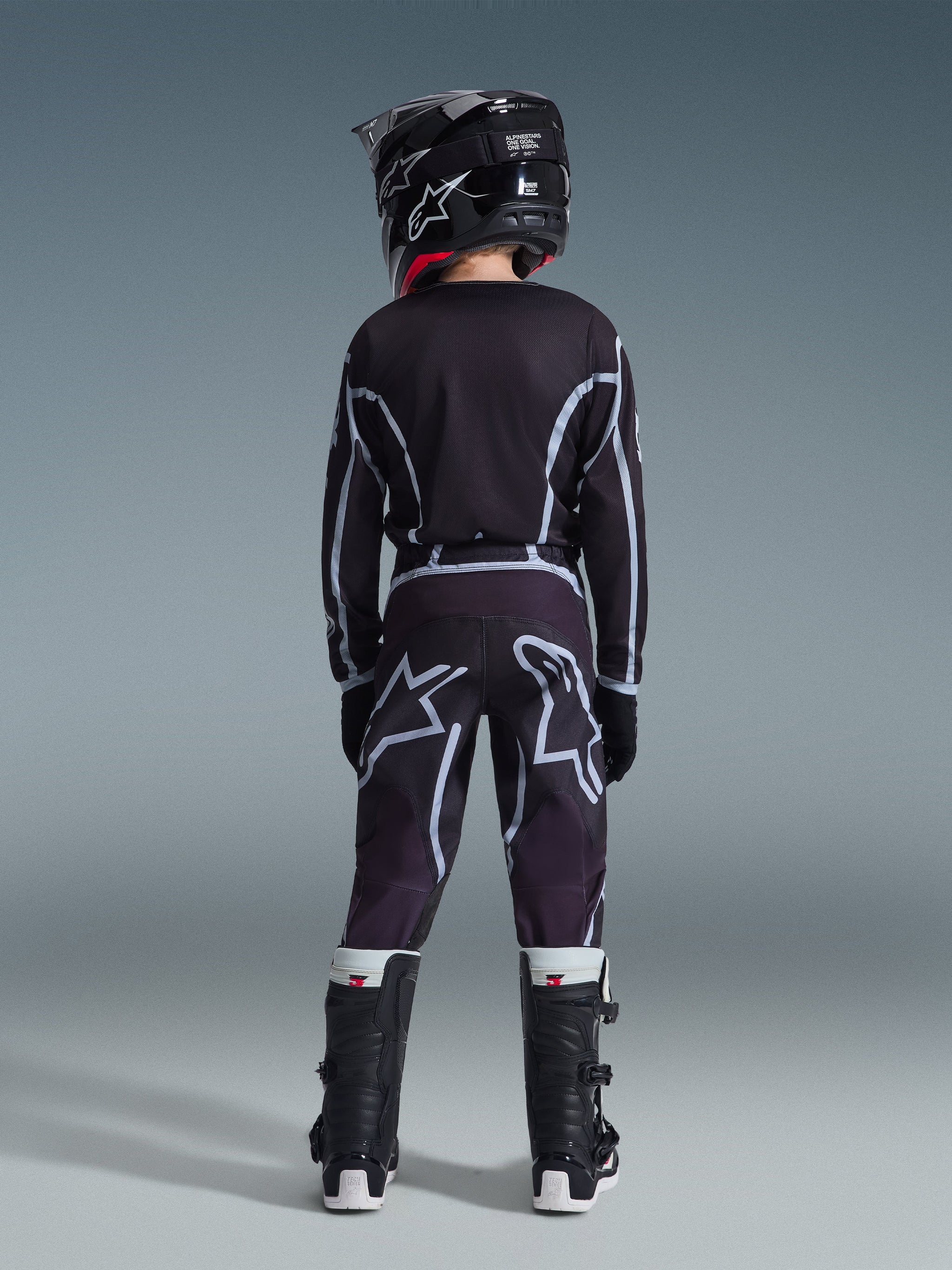 Jovem piloto de motocross visto de costas a usar a camisola 2026 Youth Fluid Apex em preto e cinzento, calças de motocross a combinar com grandes grafismos do logótipo em branco, capacete preto e botas Tech Series pretas e brancas, conjunto completo de equipamento de performance para condução todo-o-terreno.