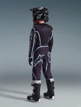 Alpinestars 2026 Youth Fluid Apex Jersey, camisola de motocross, preta e cinzenta, usada com calças Youth Fluid Grid a condizer, capacete preto e botas pretas e brancas, vista traseira mostrando o equipamento completo de corrida todo-o-terreno para jovens pilotos