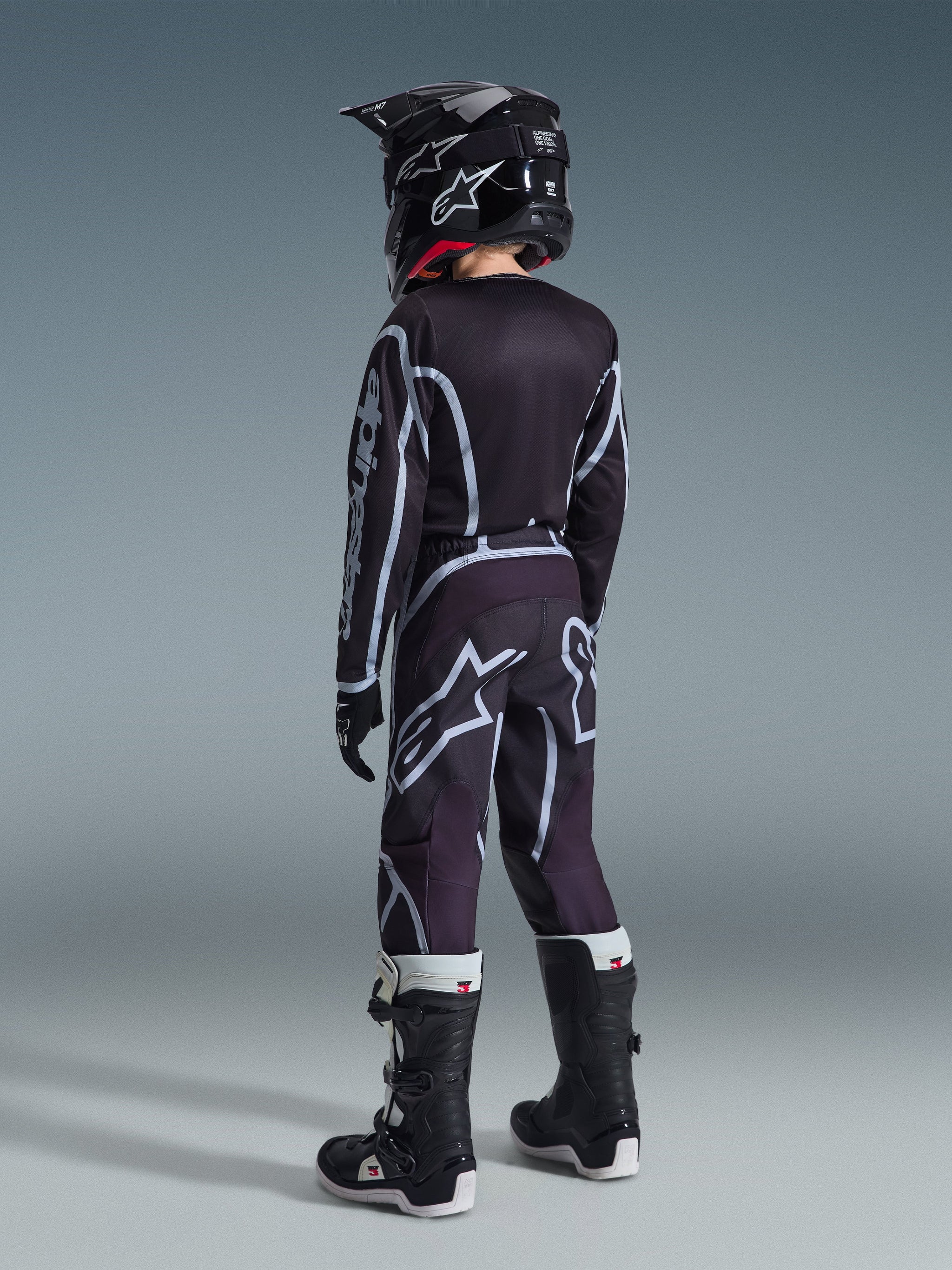 Alpinestars 2026 Youth Fluid Apex Jersey, camisola de motocross, preta e cinzenta, usada com calças Youth Fluid Grid a condizer, capacete preto e botas pretas e brancas, vista traseira mostrando o equipamento completo de corrida todo-o-terreno para jovens pilotos