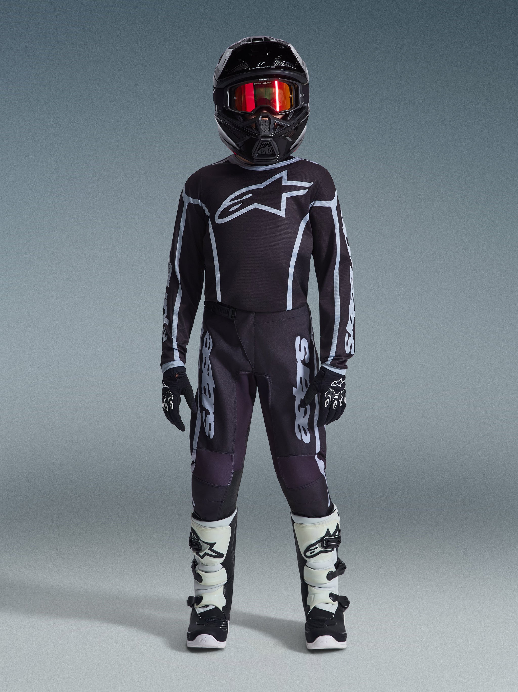 Jovem piloto de motocross a usar a Camisola Alpinestars 2026 Youth Fluid Apex em preto e cinzento, coordenada com calças a condizer, capacete preto com óculos iridescentes vermelhos, luvas pretas e brancas e botas de motocross, conjunto completo de equipamento para corridas off-road juvenis