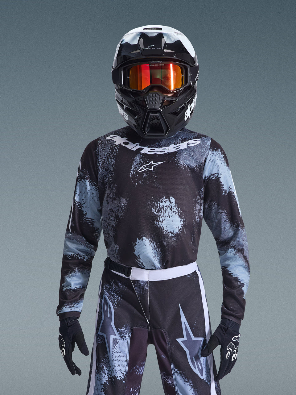 Alpinestars 2026 Youth Fluid Lahnd Jersey, camisola de motocross, Iron Camo, usada por um jovem piloto com calças a condizer, capacete preto e branco com óculos cor de laranja e luvas pretas, com um design salpicado em cinzento e preto e logótipo branco, equipamento de corrida off-road completo para jovens pilotos.