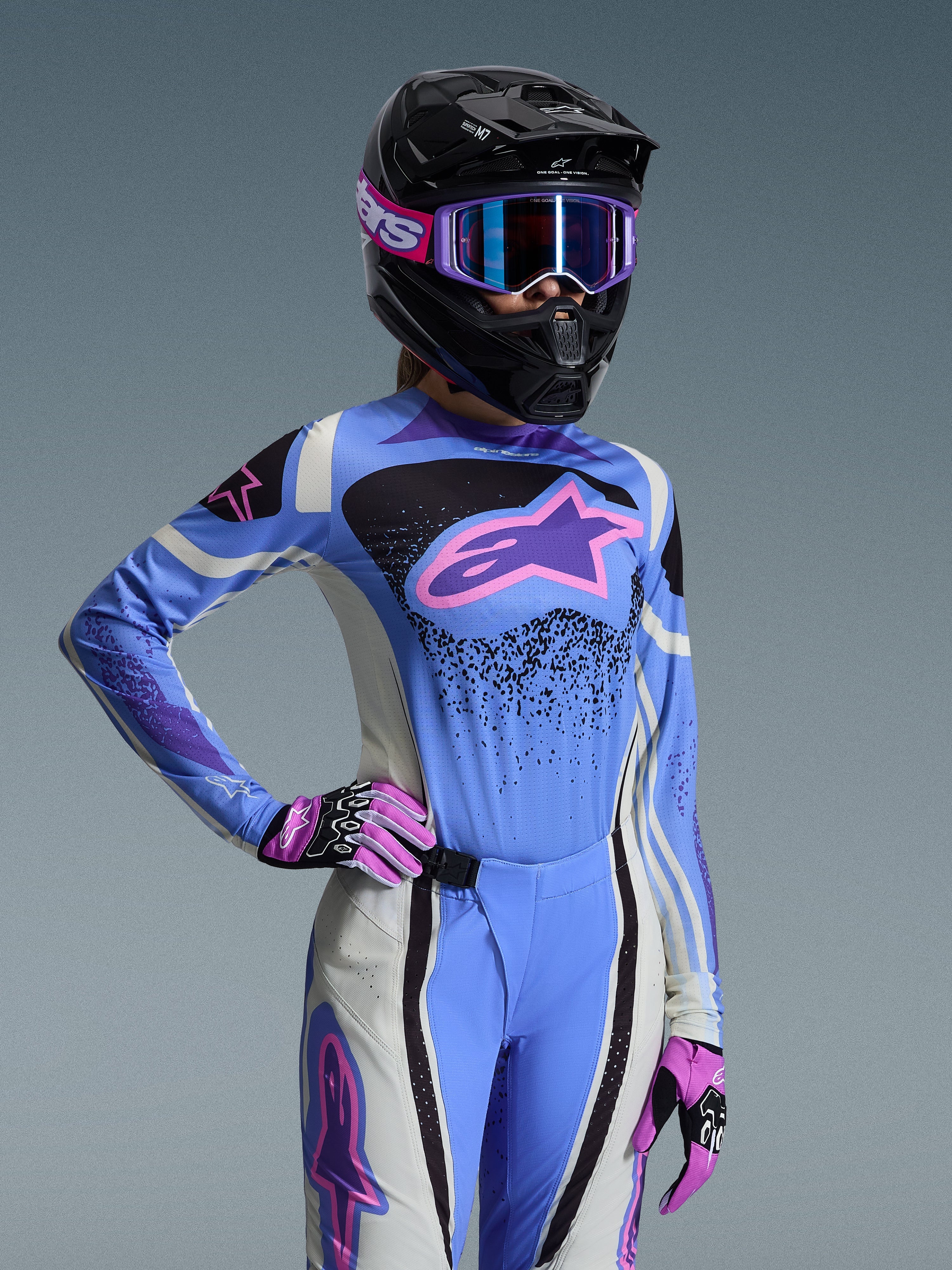 Piloto de motocross feminina com o conjunto de equipamento Alpinestars Stella Techstar Nomur 2026, camisola azul clara com detalhes em cor-de-rosa e roxo, calças em azul e branco a condizer, capacete integral preto com óculos roxos e luvas cor-de-rosa e pretas, equipamento profissional de corrida todo-o-terreno para mulher
