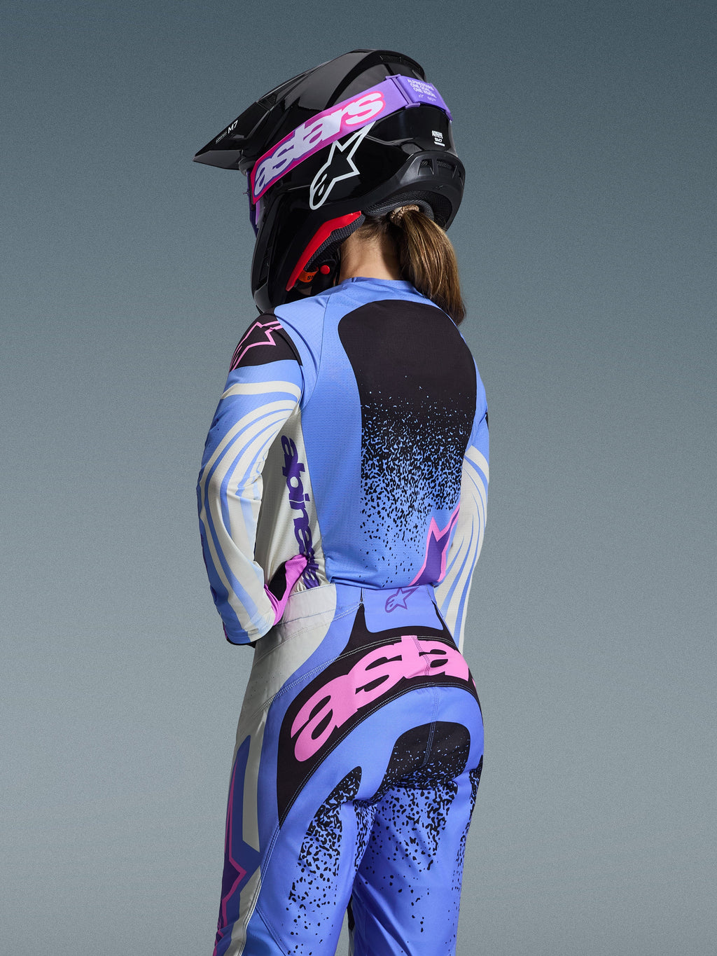 Vista traseira de uma piloto de motocross a usar o conjunto de equipamento Alpinestars 2026 Stella Techstar Nomur, azul claro com detalhes em cor-de-rosa e preto, camisola e calças a condizer, capacete preto com óculos cor-de-rosa, corte feminino ergonómico para competição off-road.