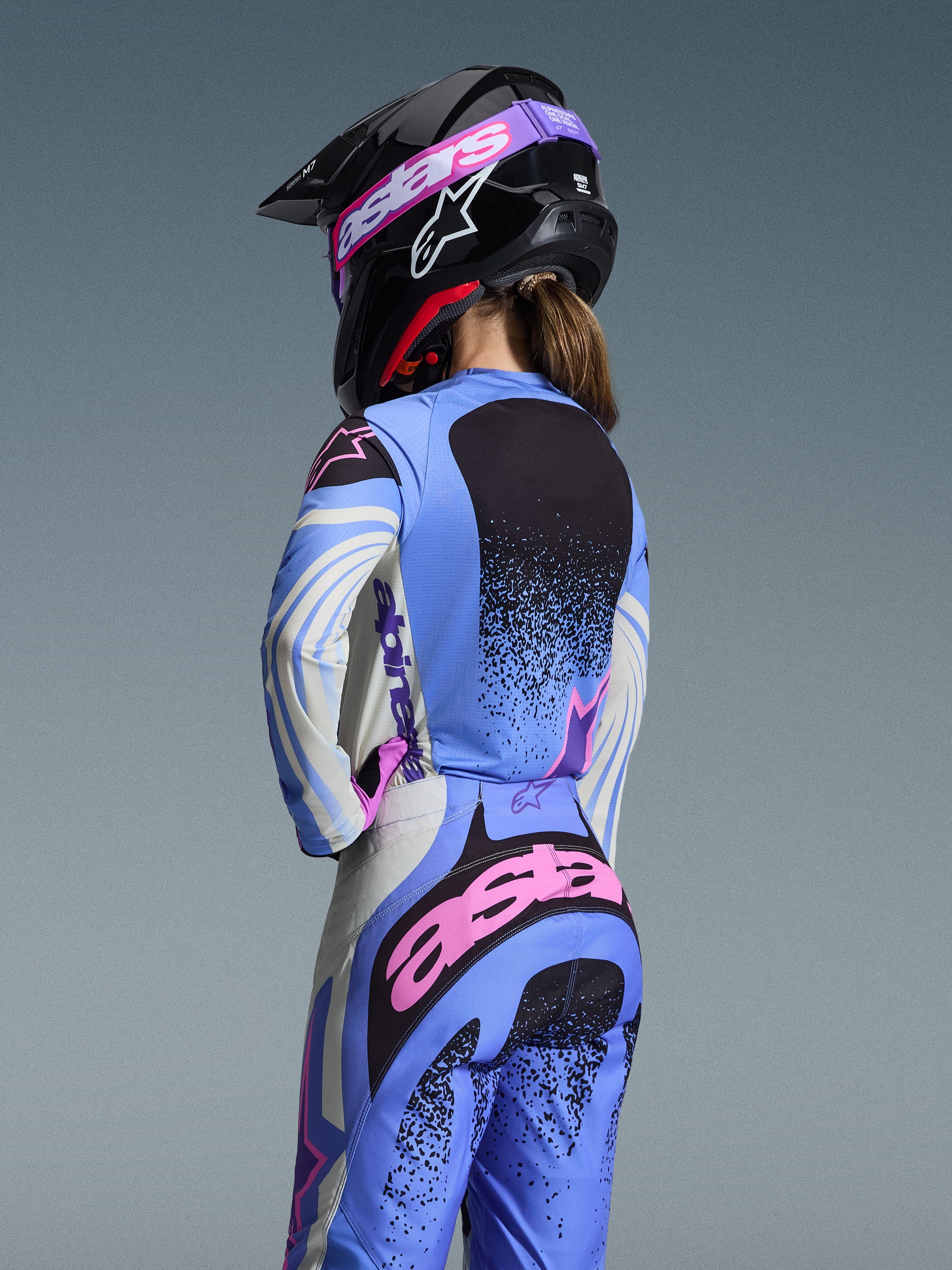 Vista traseira de uma piloto de motocross a usar o conjunto de equipamento Alpinestars 2026 Stella Techstar Nomur, azul claro com detalhes em cor-de-rosa e preto, camisola e calças a condizer, capacete preto com óculos cor-de-rosa, corte feminino ergonómico para competição off-road.