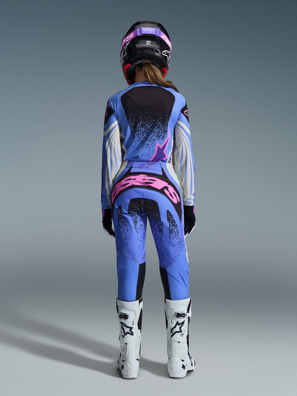 Vista traseira de uma piloto de motocross a usar o conjunto Alpinestars 2026 Stella Techstar Nomur, camisola e calças azuis com detalhes em cor-de-rosa e branco, logótipo proeminente em cor-de-rosa na parte de trás das calças, botas de motocross brancas e capacete preto e cor-de-rosa a condizer.