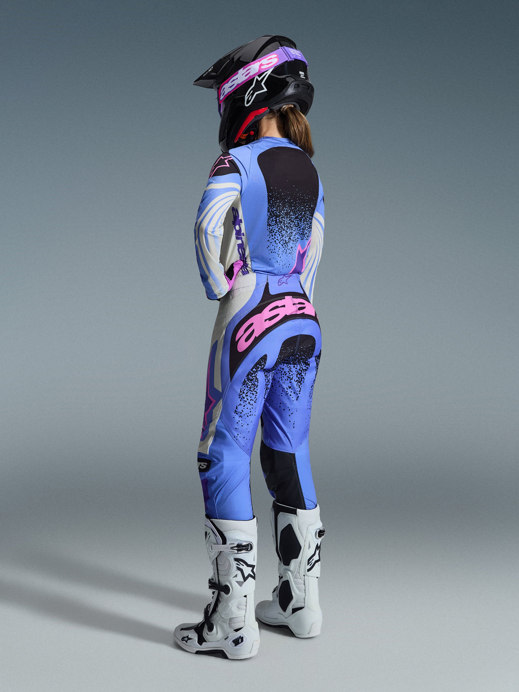 Alpinestars 2026 Stella Techstar Nomur Gear Set, vista traseira de uma piloto de motocross a usar um conjunto de camisola e calças em azul claro, branco e rosa com detalhes salpicados a preto, combinado com um capacete preto e botas brancas, equipamento de corrida profissional para mulher