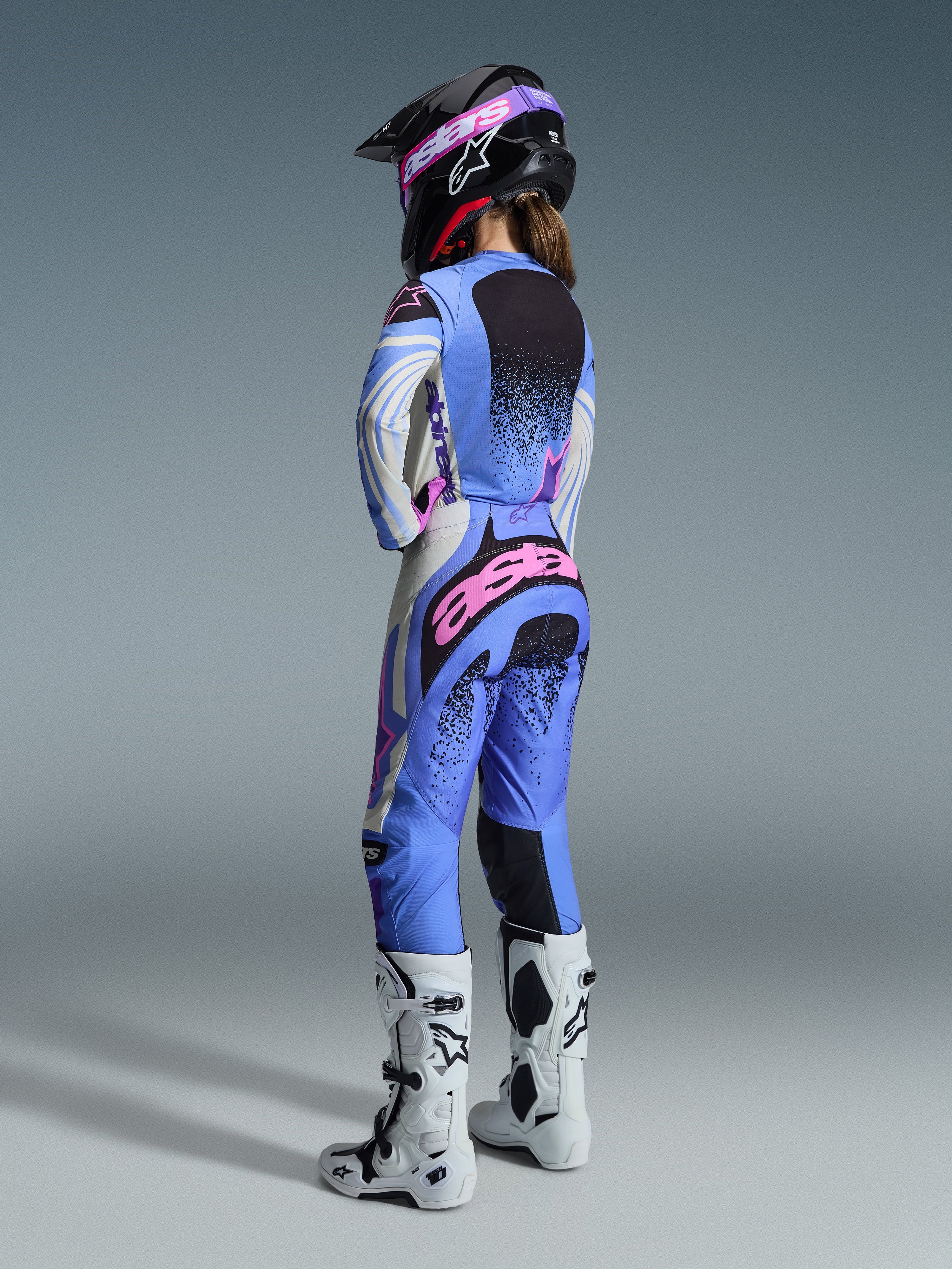 Alpinestars 2026 Stella Techstar Nomur Gear Set, vista traseira de uma piloto de motocross a usar um conjunto de camisola e calças em azul claro, branco e rosa com detalhes salpicados a preto, combinado com um capacete preto e botas brancas, equipamento de corrida profissional para mulher