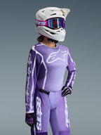 Piloto de motocross com o conjunto de equipamento Alpinestars 2026 Stella Fluid Apex para mulher, camisola roxa e branca com logótipos Alpinestars, calças roxas a condizer, capacete branco com óculos roxos e luvas pretas e roxas, equipamento profissional de corrida todo-o-terreno