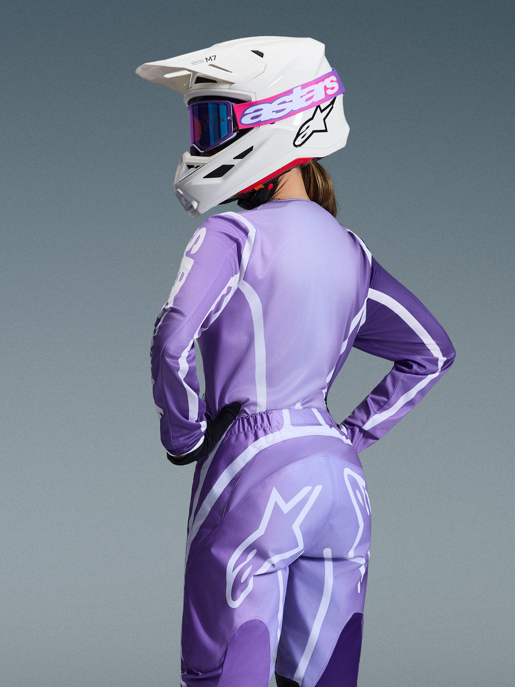 Piloto de motocross a usar o Conjunto de Equipamento Alpinestars 2026 Stella Fluid Apex, camisola e calças roxas e lavanda com grafismos brancos, capacete branco com óculos cor-de-rosa e roxos, ajuste específico para mulher, equipamento coordenado para corridas de todo-o-terreno