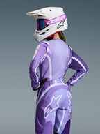 Piloto de motocross a usar o Conjunto de Equipamento Alpinestars 2026 Stella Fluid Apex, camisola e calças roxas e lavanda com grafismos brancos, capacete branco com óculos cor-de-rosa e roxos, ajuste específico para mulher, equipamento coordenado para corridas de todo-o-terreno