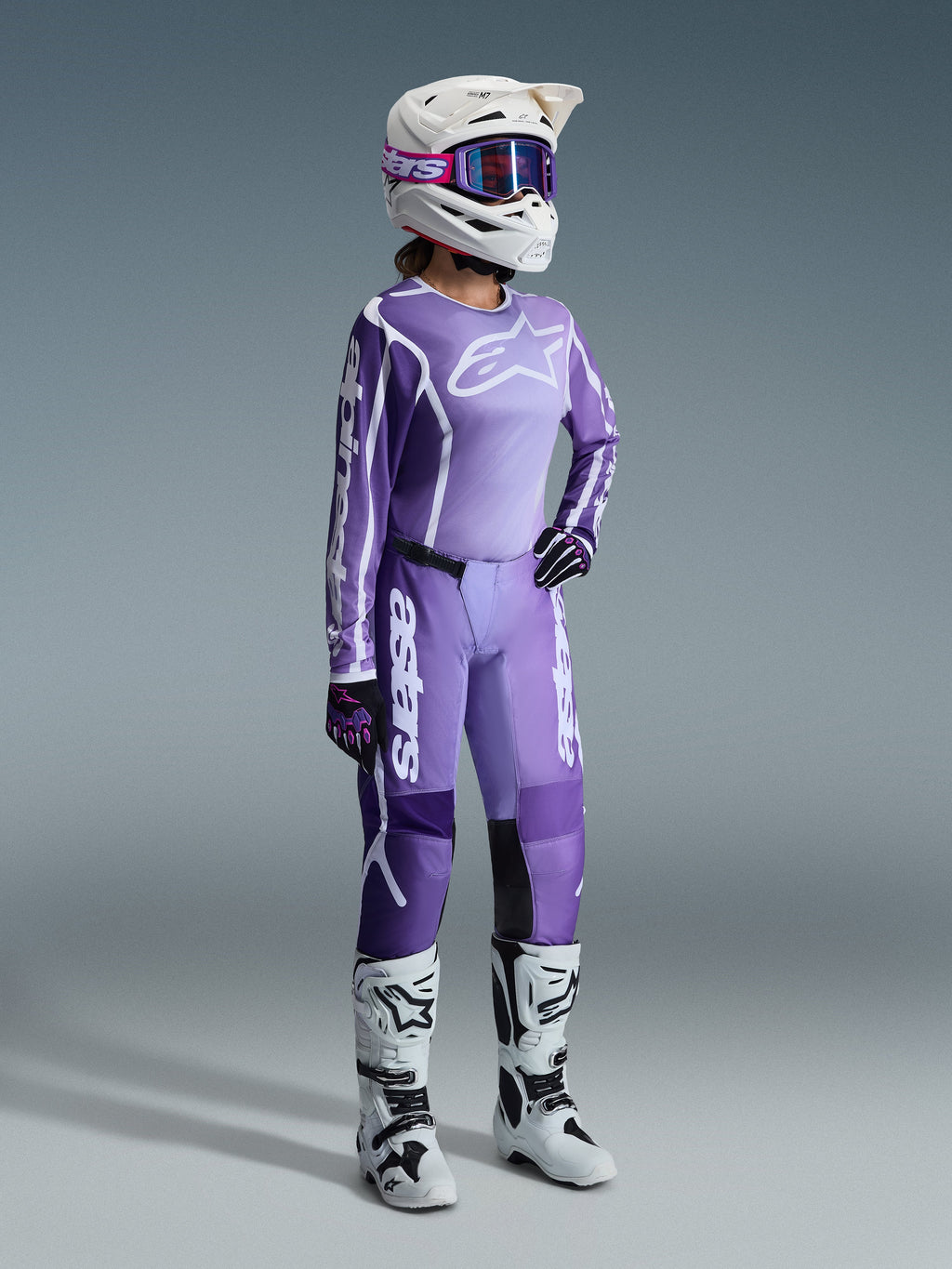 Conjunto de Equipamento Alpinestars Stella Fluid Apex 2026, roxo e lavanda, piloto feminina de motocross a usar camisola e calças a condizer, capacete branco com óculos de proteção roxos, botas brancas e pretas, luvas pretas e roxas, equipamento completo de alta performance para competição todo-o-terreno feminina