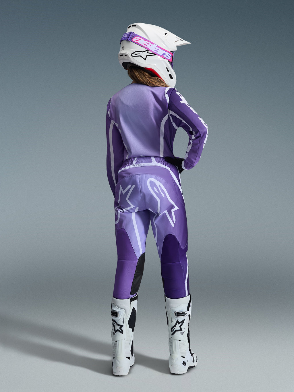 Conjunto de Equipamento Alpinestars Stella Fluid Apex 2026, equipamento de motocross para mulher, roxo e lavanda com detalhes brancos, pilota vista de costas com camisola e calças a condizer, capacete branco com óculos cor-de-rosa e botas brancas, conjunto de corrida atlético coordenado