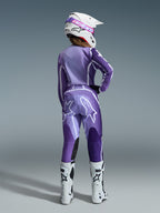 Conjunto de Equipamento Alpinestars Stella Fluid Apex 2026, equipamento de motocross para mulher, roxo e lavanda com detalhes brancos, pilota vista de costas com camisola e calças a condizer, capacete branco com óculos cor-de-rosa e botas brancas, conjunto de corrida atlético coordenado