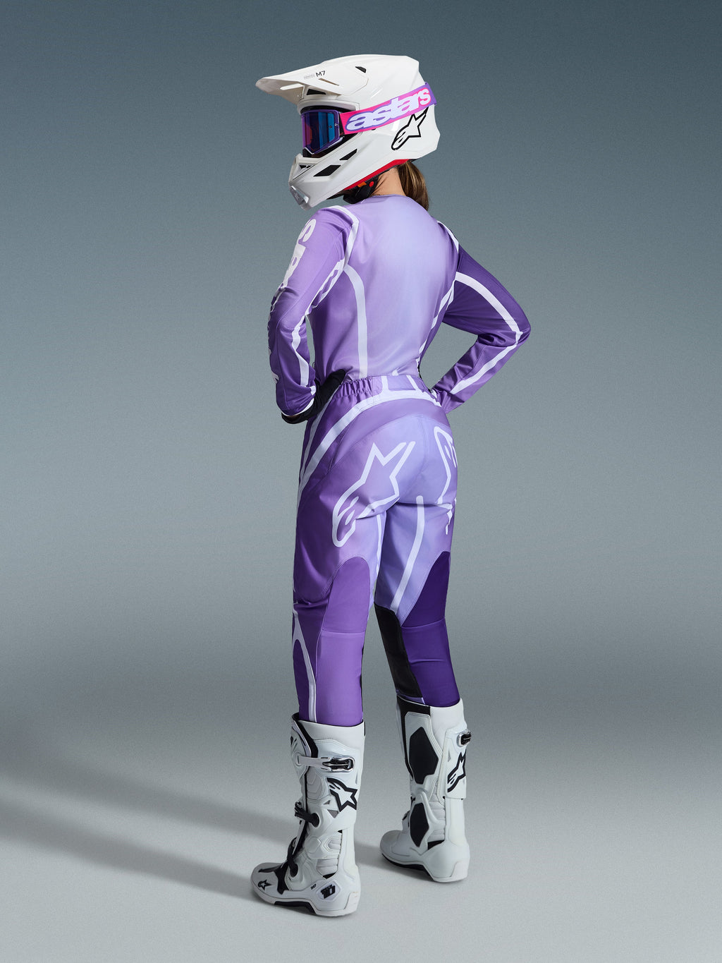 Piloto feminina de motocross com o Conjunto Stella Fluid Apex Feminino 2026, camisola e calças roxas e lavanda com detalhes brancos, capacete branco com óculos cor-de-rosa, botas brancas e pretas, conjunto coordenado para corridas todo-o-terreno