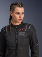Casaco Alpinestars Stella ST-2 Air, casaco de motociclismo para mulher em preto, com painéis de malha para fluxo de ar, marca "Alpinestars Exploration Equipment" com coordenadas e logótipo vermelho no peito, usado por uma modelo com cabelo entrançado.