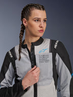 Woman wearing the Alpinestars Stella ST-2 Air Jacket in Ice Gray and Black, close-up portrait, featuring mesh panels for ventilation, 'ALPINESTARS EXPLORATION EQUIPMENT' branding on the chest with a blue logo, modern adventure motorcycle jacket