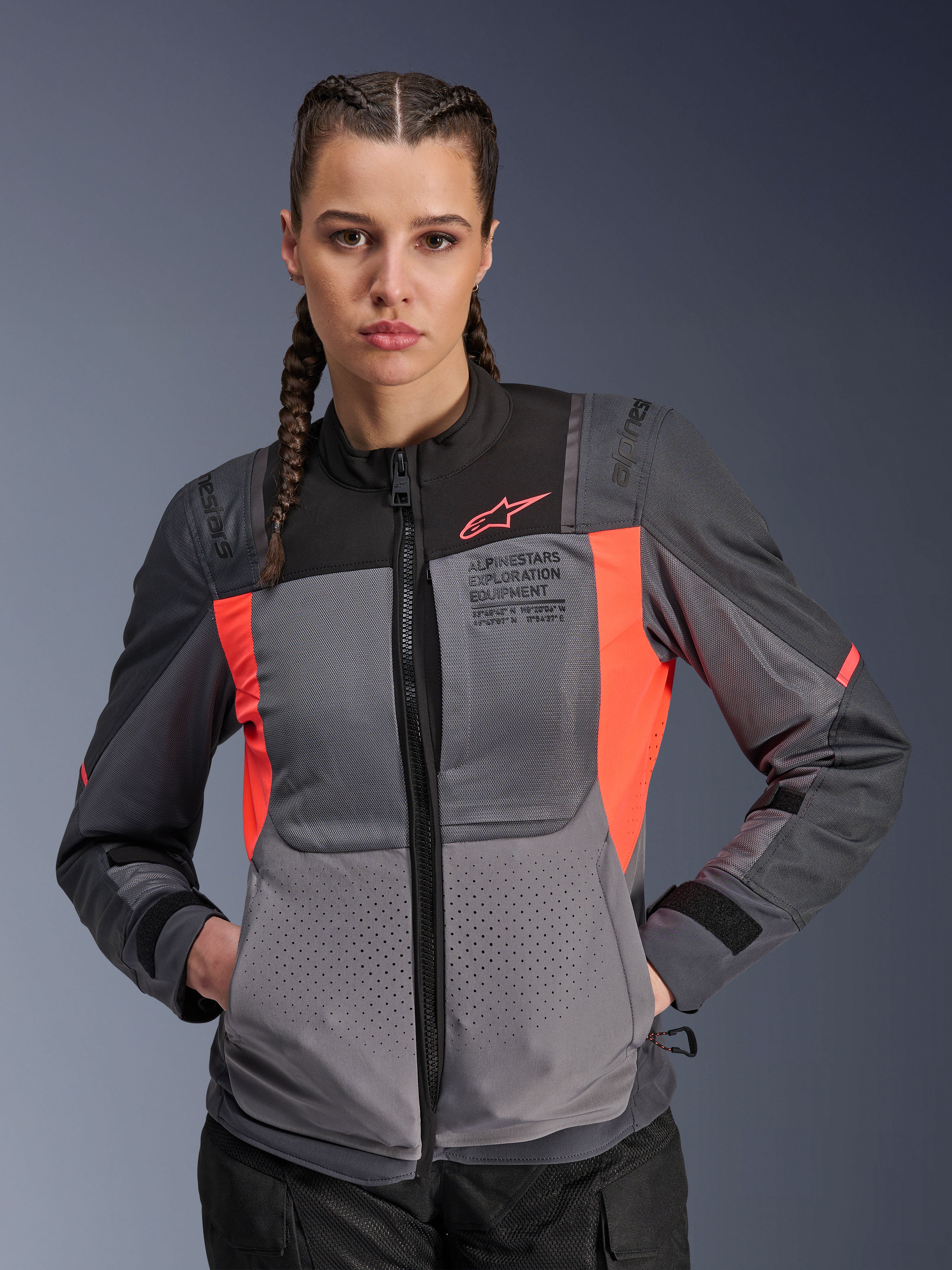 Casaco Alpinestars Stella ST-2 Air, casaco de motociclismo feminino, cinzento escuro e coral flúor, usado por uma modelo com tranças, com painéis de malha tecnológica para fluxo de ar, perfurações a laser e a marca "Alpinestars Exploration Equipment" no peito, equipamento de touring de verão