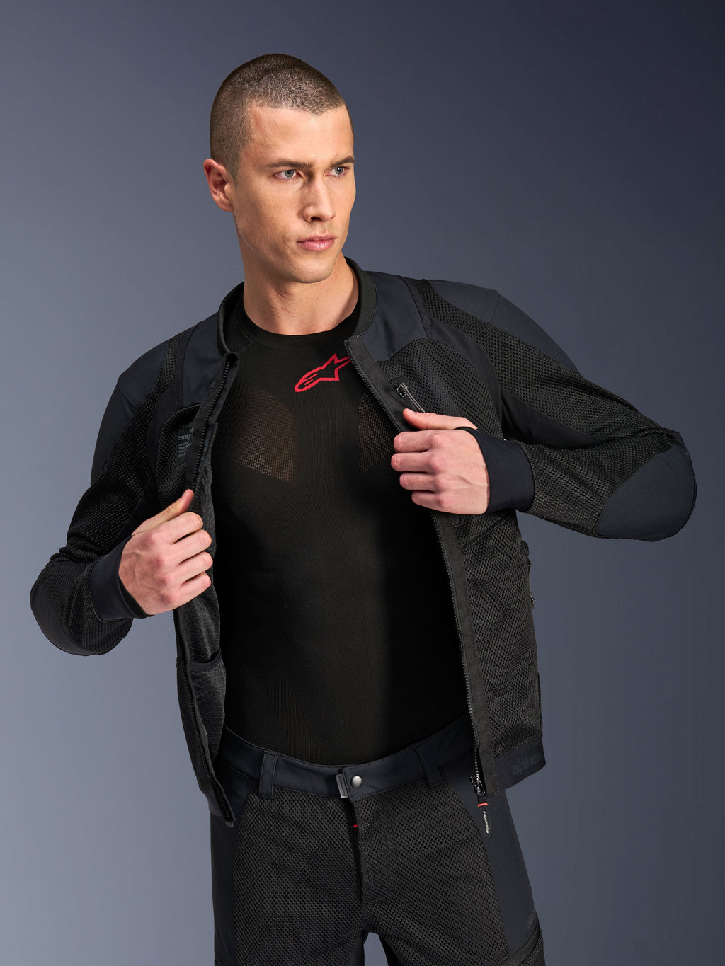 Alpinestars Troop-Air Pro Jacket, preto, casaco de ar blindado técnico premium com construção em malha, usado aberto sobre uma camada de base preta com um logótipo vermelho, combinado com calças de motociclismo em malha a condizer, estilo de condução urbano contemporâneo