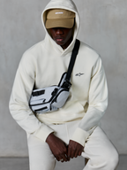 Alpinestars Essentials Ageless Hoodie, streetwear casual, cor Off White, pequeno logótipo preto bordado no peito, capuz usado sobre um boné bege com a marca ASTARS, combinado com uma mala de tiracolo cinzenta clara, vestuário lifestyle premium