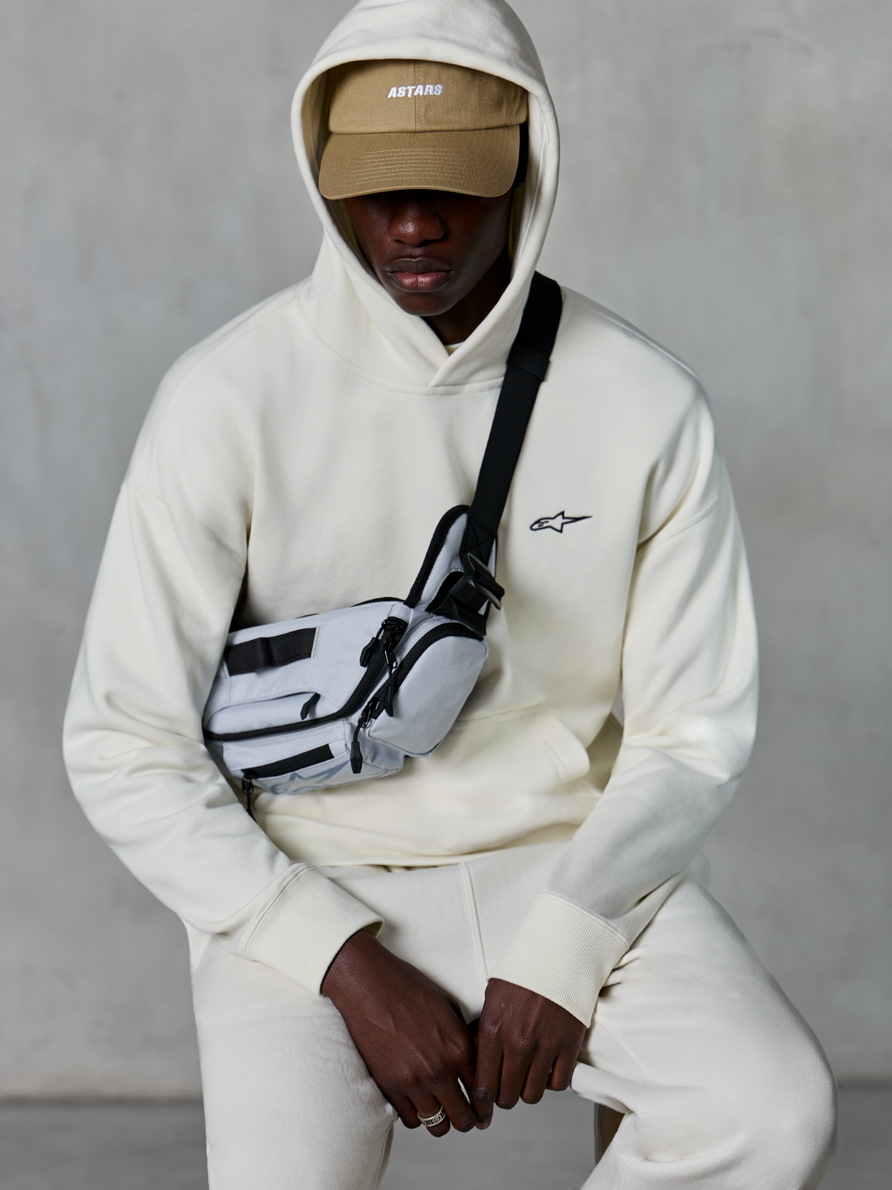 Alpinestars Essentials Ageless Hoodie, streetwear casual, cor Off White, pequeno logótipo preto bordado no peito, capuz usado sobre um boné bege com a marca ASTARS, combinado com uma mala de tiracolo cinzenta clara, vestuário lifestyle premium