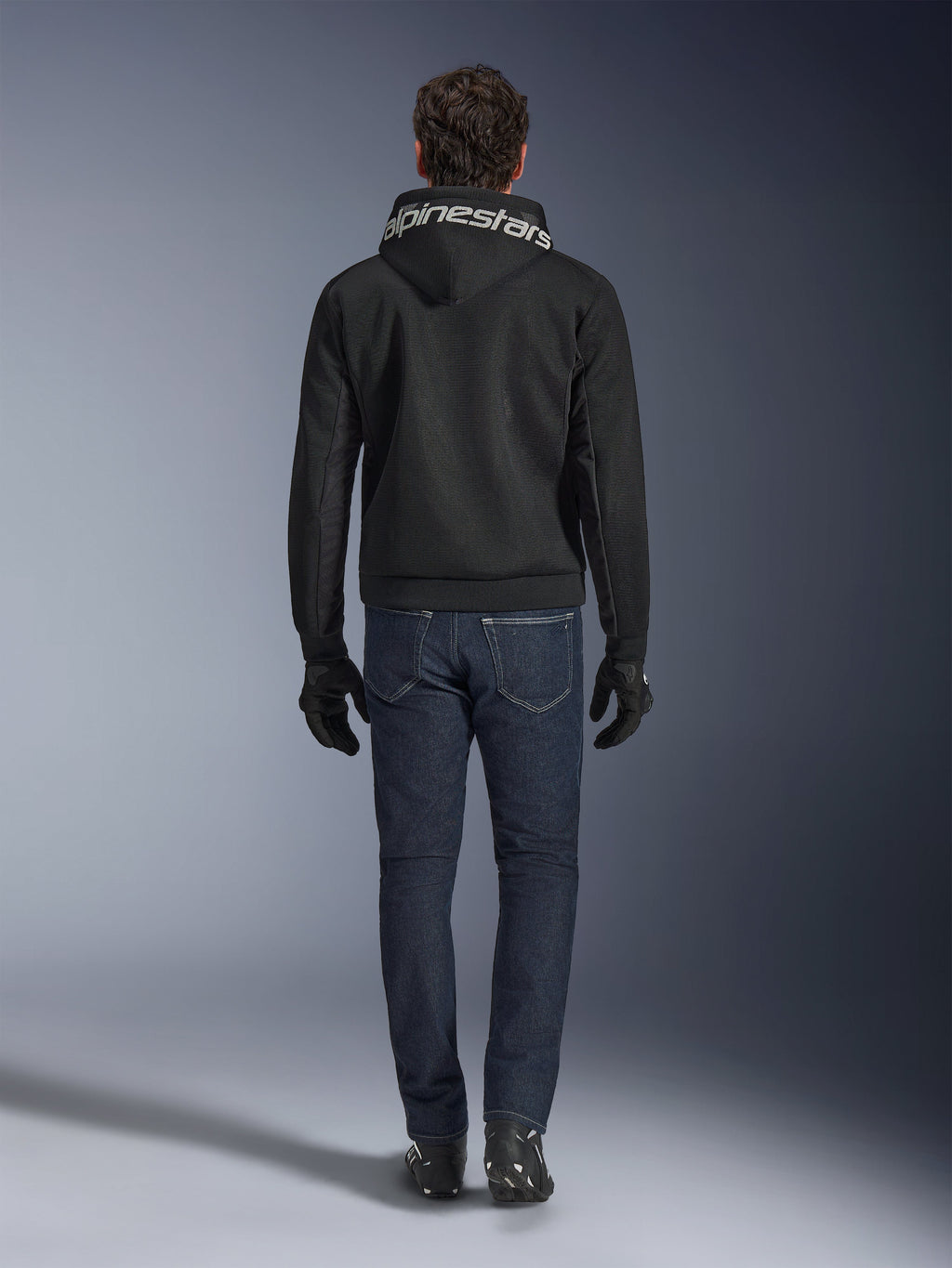 Alpinestars Chrome Superairflow Hoodie, Black motorcycle hoodie with white branding on the hood, viewed from the back, worn with dark blue jeans, black gloves, and black and white boots, urban sport riding gear
