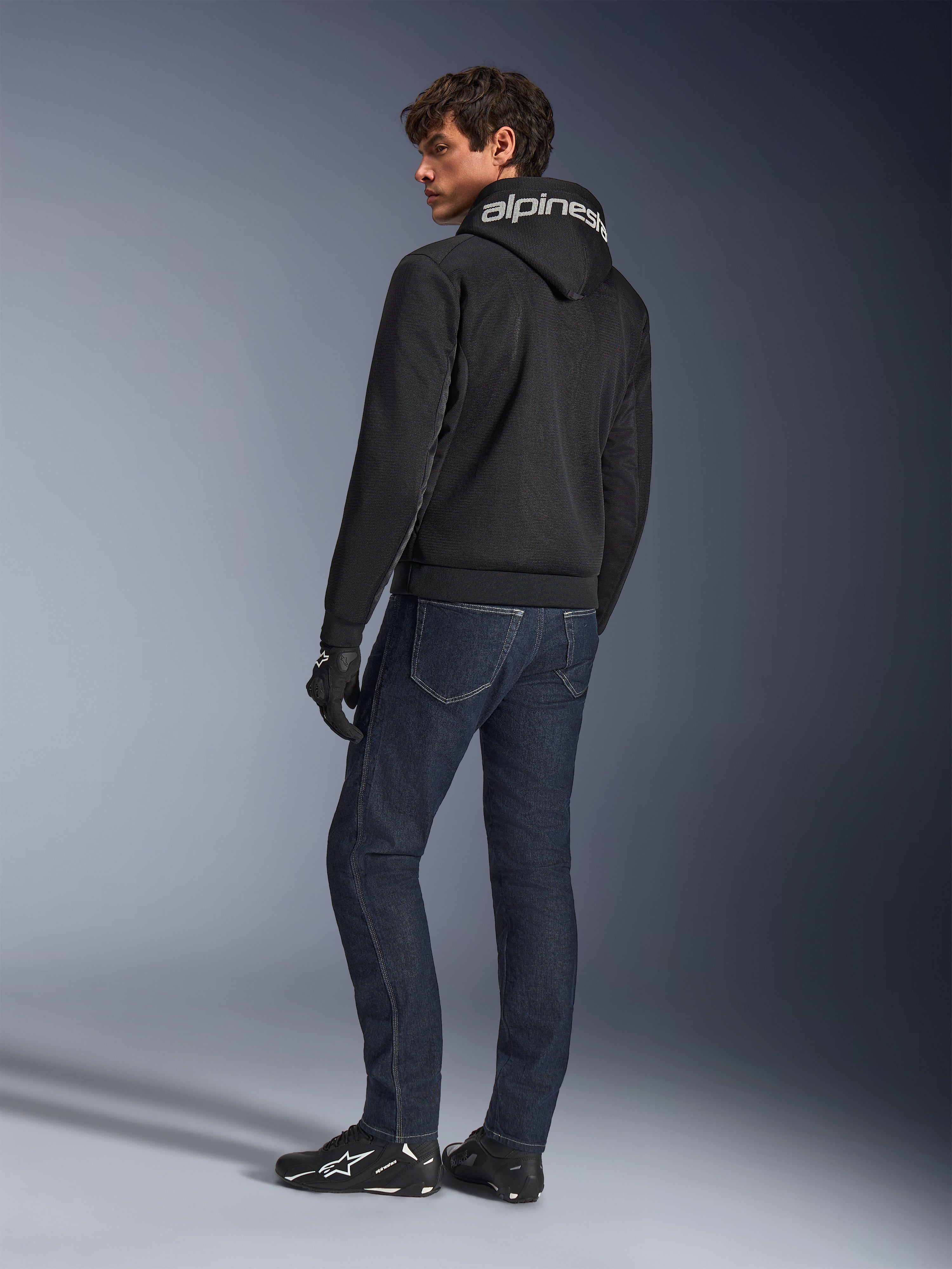 Alpinestars Chrome Superairflow Hoodie, Black, back view on male model, featuring white Alpinestars branding on the hood, worn with dark denim jeans and black gloves, urban motorcycle gear design