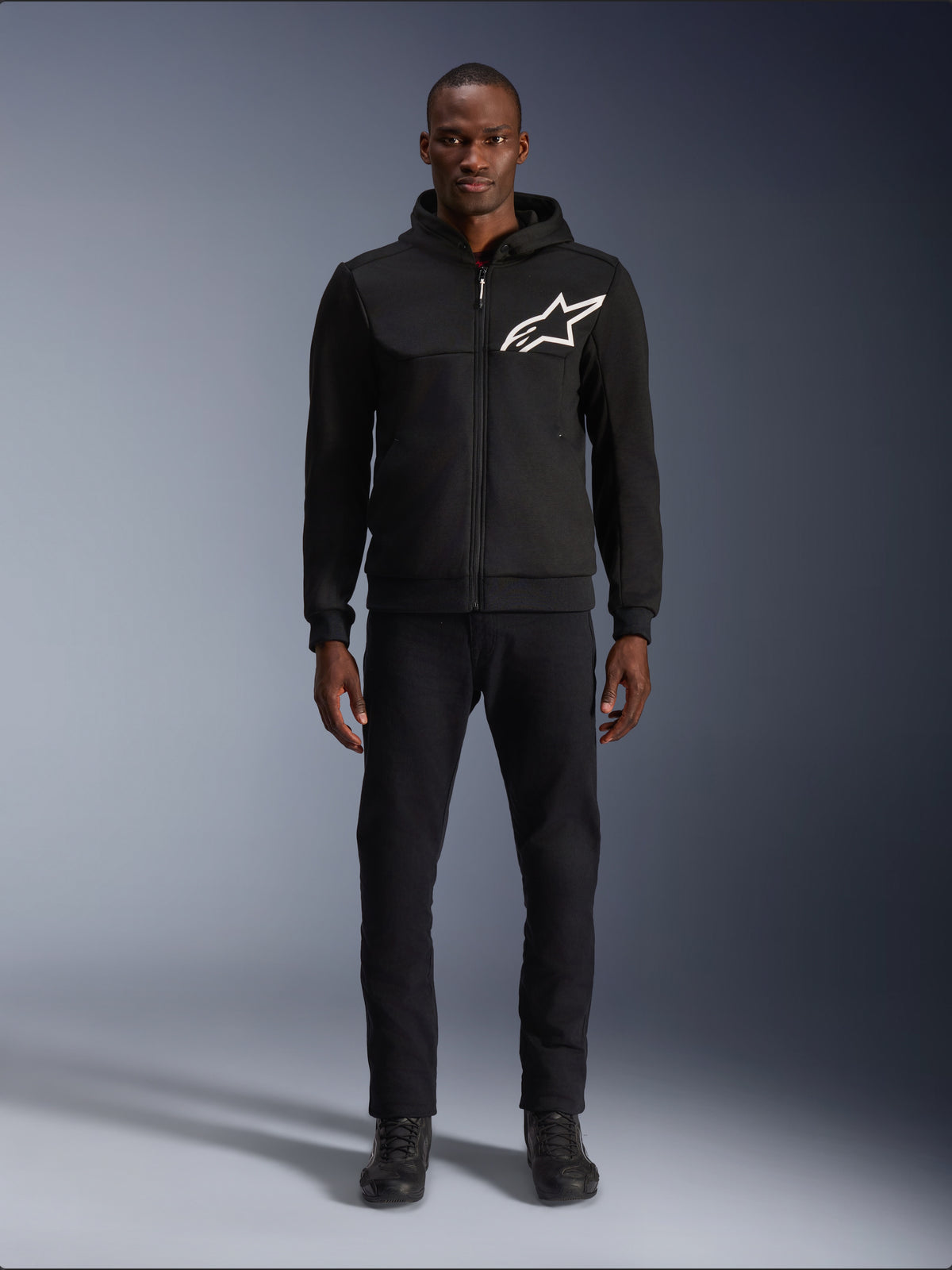 Alpinestars Chrome V2 Sport Hoodie, Black and White motorcycle hoodie, featuring a large white Alpinestars logo on the chest, zip-up front and hood, worn by a model standing with black pants and boots, urban-style protective riding gear.