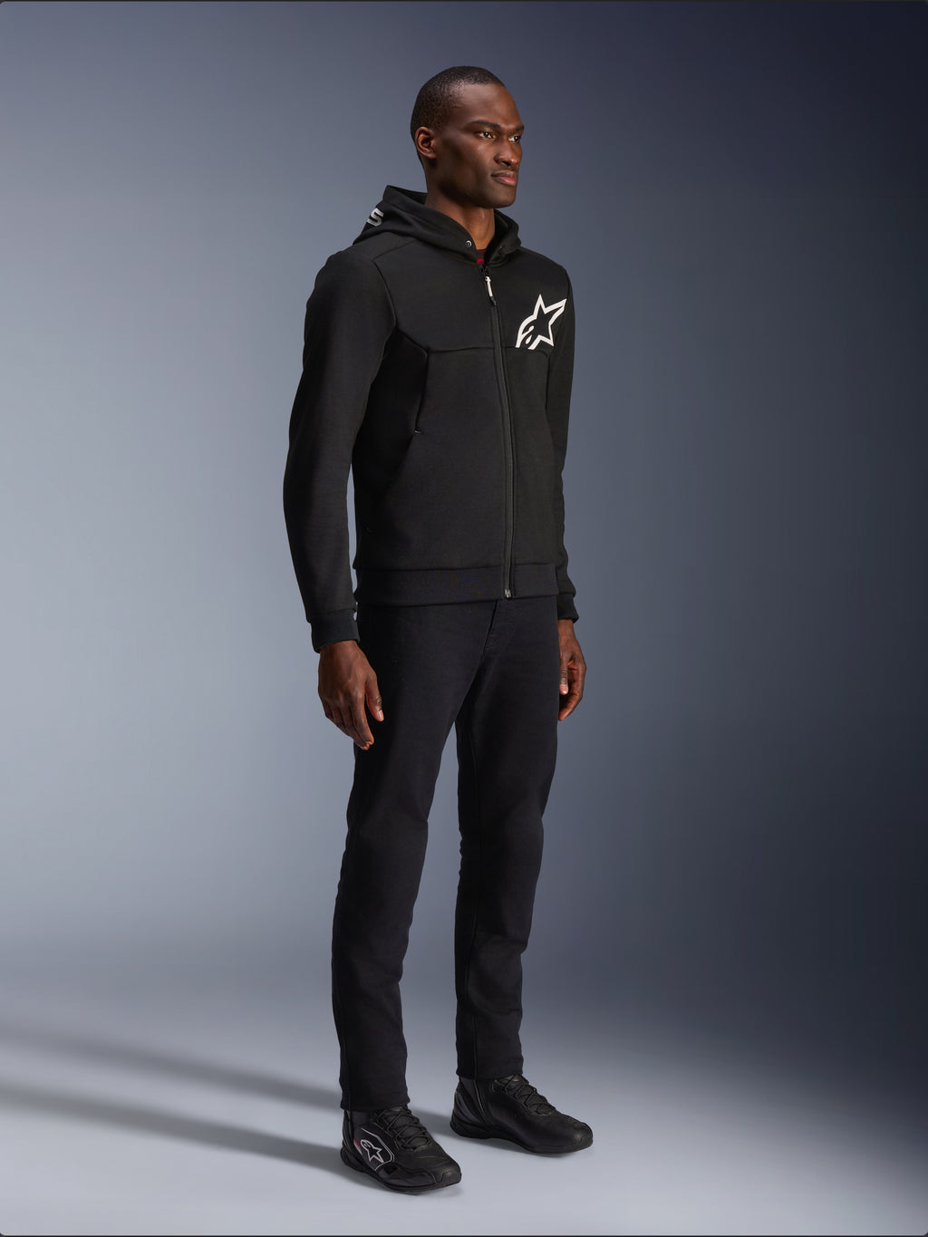 Alpinestars Chrome V2 Sport Hoodie, black motorcycle hoodie with white star logo, worn by a male model with matching black pants and riding sneakers, urban lifestyle riding gear