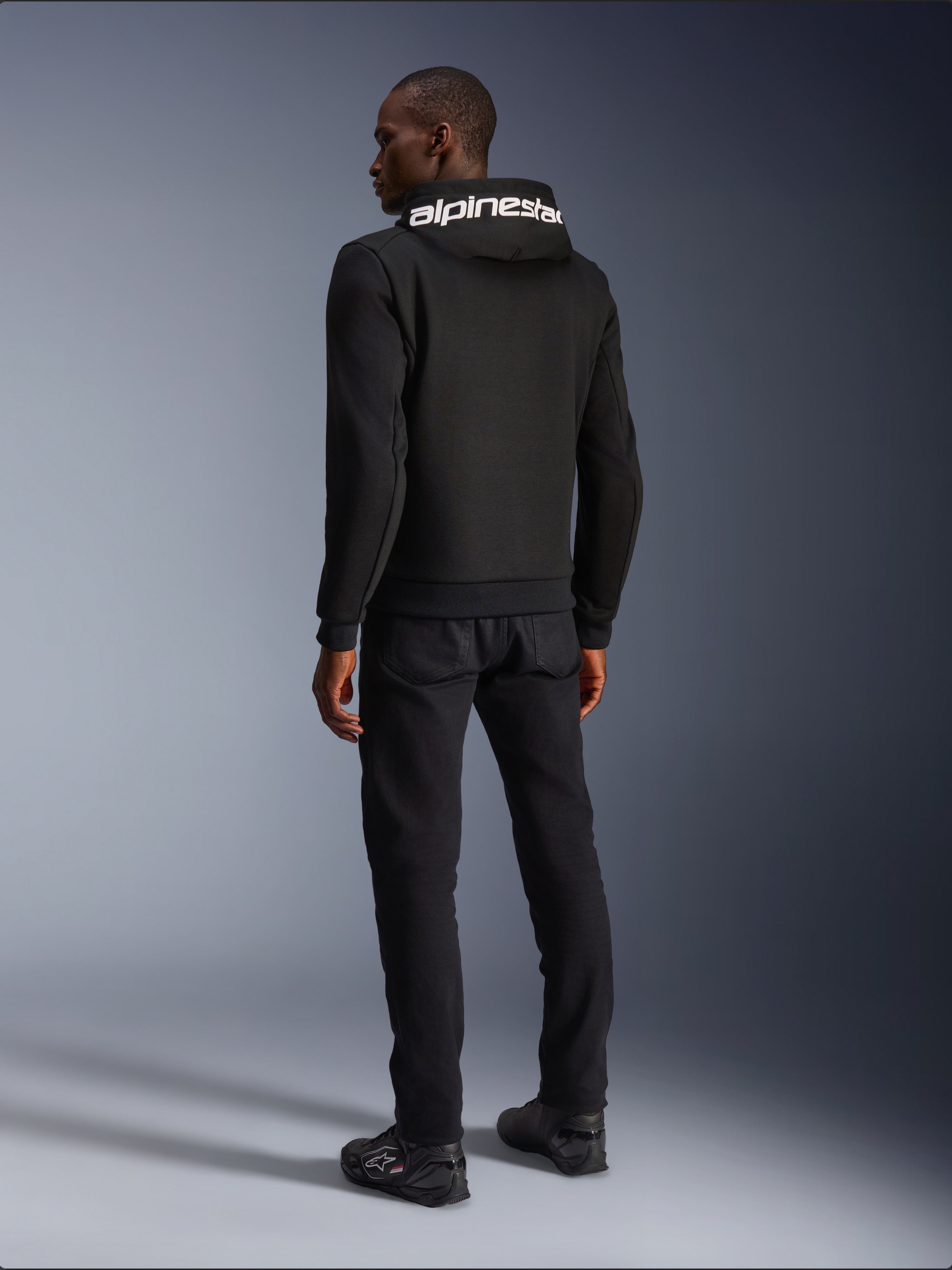 Alpinestars Chrome V2 Sport Hoodie, Black, rear view showing white Alpinestars branding on the hood, worn with black pants and riding shoes, casual urban motorcycle apparel with an athletic silhouette