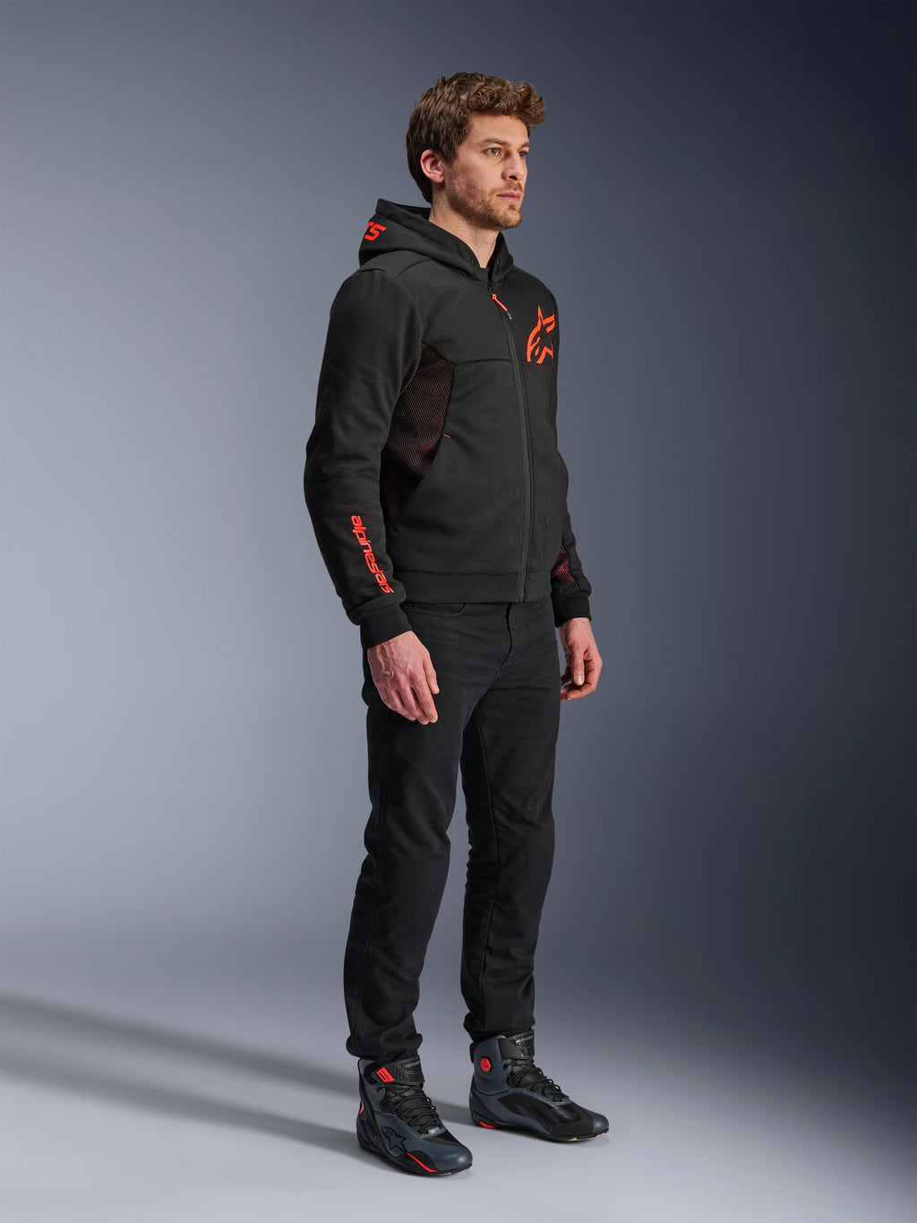 Alpinestars Chrome Air Sport Hoodie, Preto Vermelho Fluo, casaco de condução urbana com tech fleece, usado com calças pretas e botas de motociclismo, apresentando painéis de ventilação em malha e logótipos em vermelho fluorescente no peito e na manga.