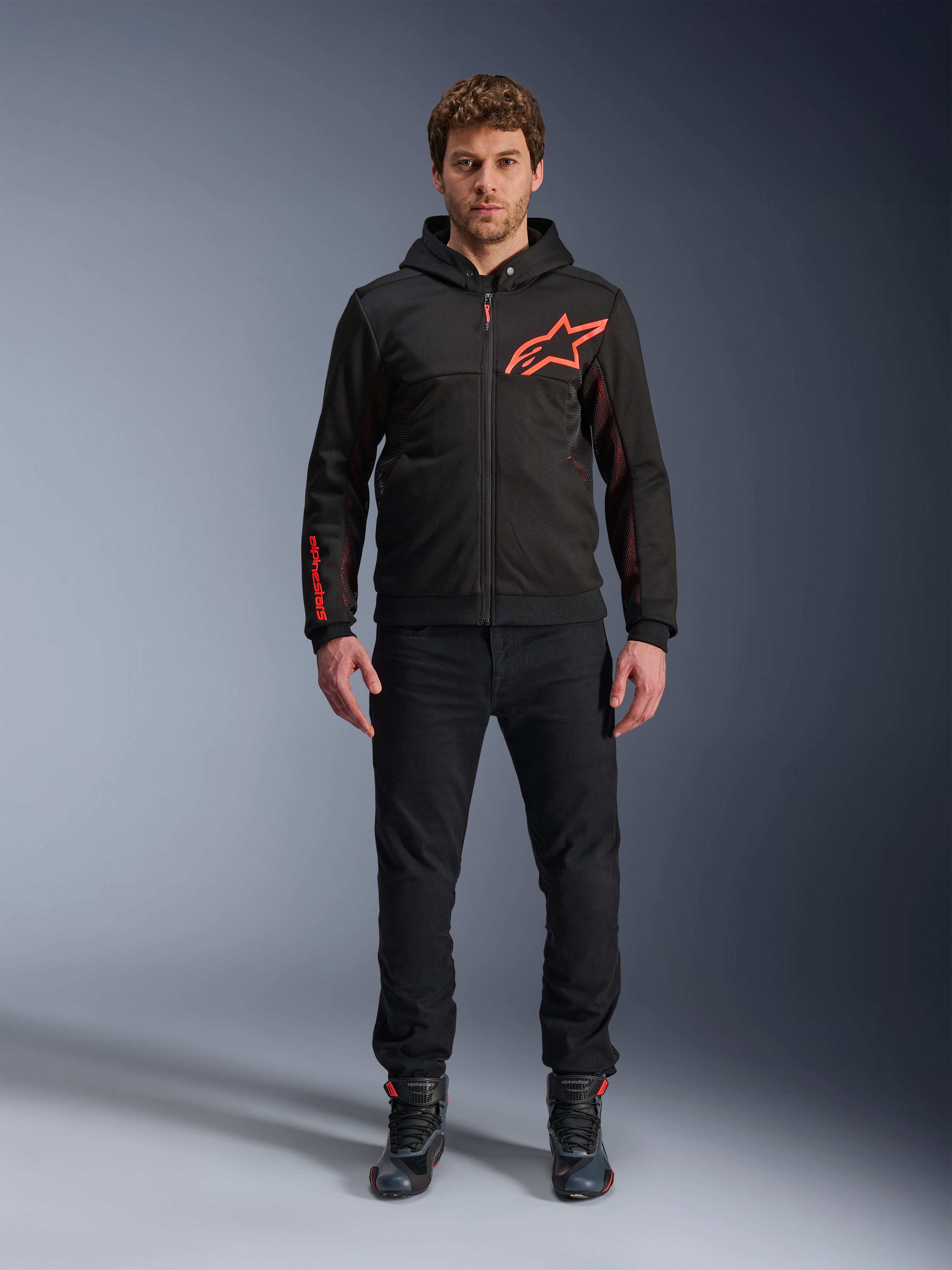 Alpinestars Chrome Air Sport Hoodie, preto e vermelho fluo, casaco de motociclismo urbano com construção em tech fleece, com um grande logótipo Astars vermelho no peito e marca na manga, vestido por um modelo com calças pretas e sapatos de condução