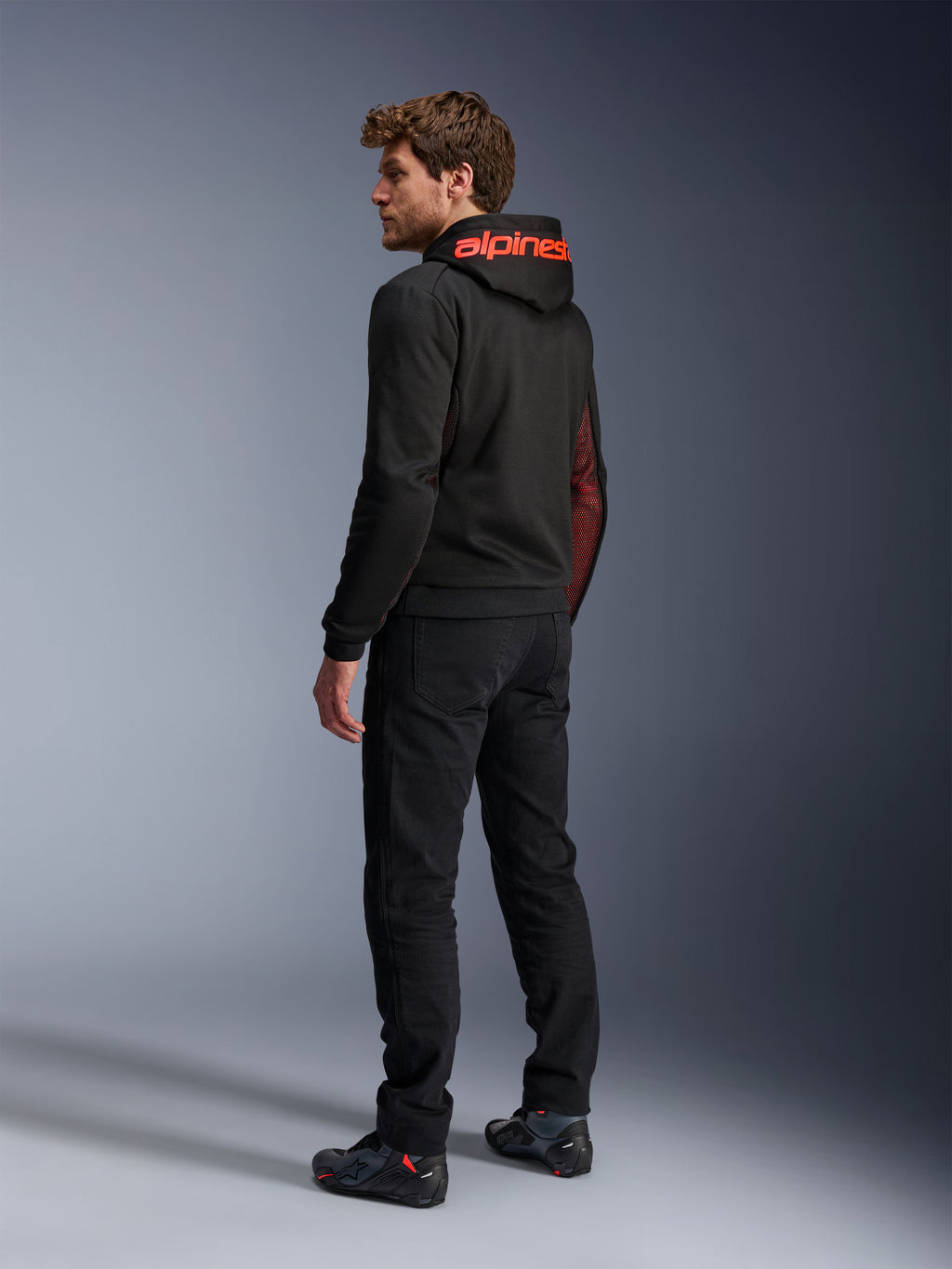 Alpinestars Chrome Air Sport Hoodie, hoodie de motociclismo urbano, preto e vermelho fluo, vista traseira mostrando a marca Alpinestars a vermelho no capuz e painéis laterais em malha, usado com calças de condução pretas e calçado técnico