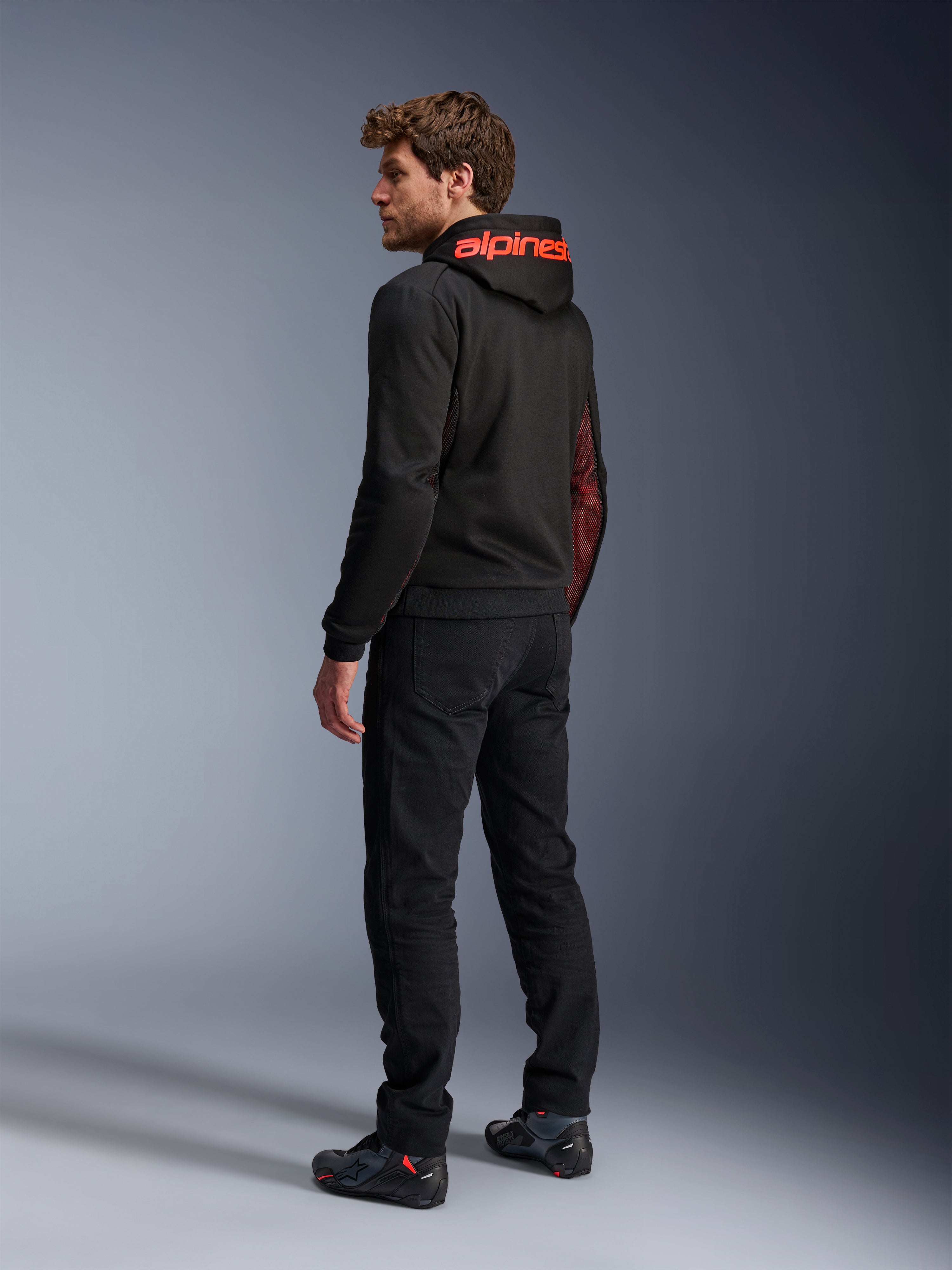 Alpinestars Chrome Air Sport Hoodie, hoodie de motociclismo urbano, preto e vermelho fluo, vista traseira mostrando a marca Alpinestars a vermelho no capuz e painéis laterais em malha, usado com calças de condução pretas e calçado técnico
