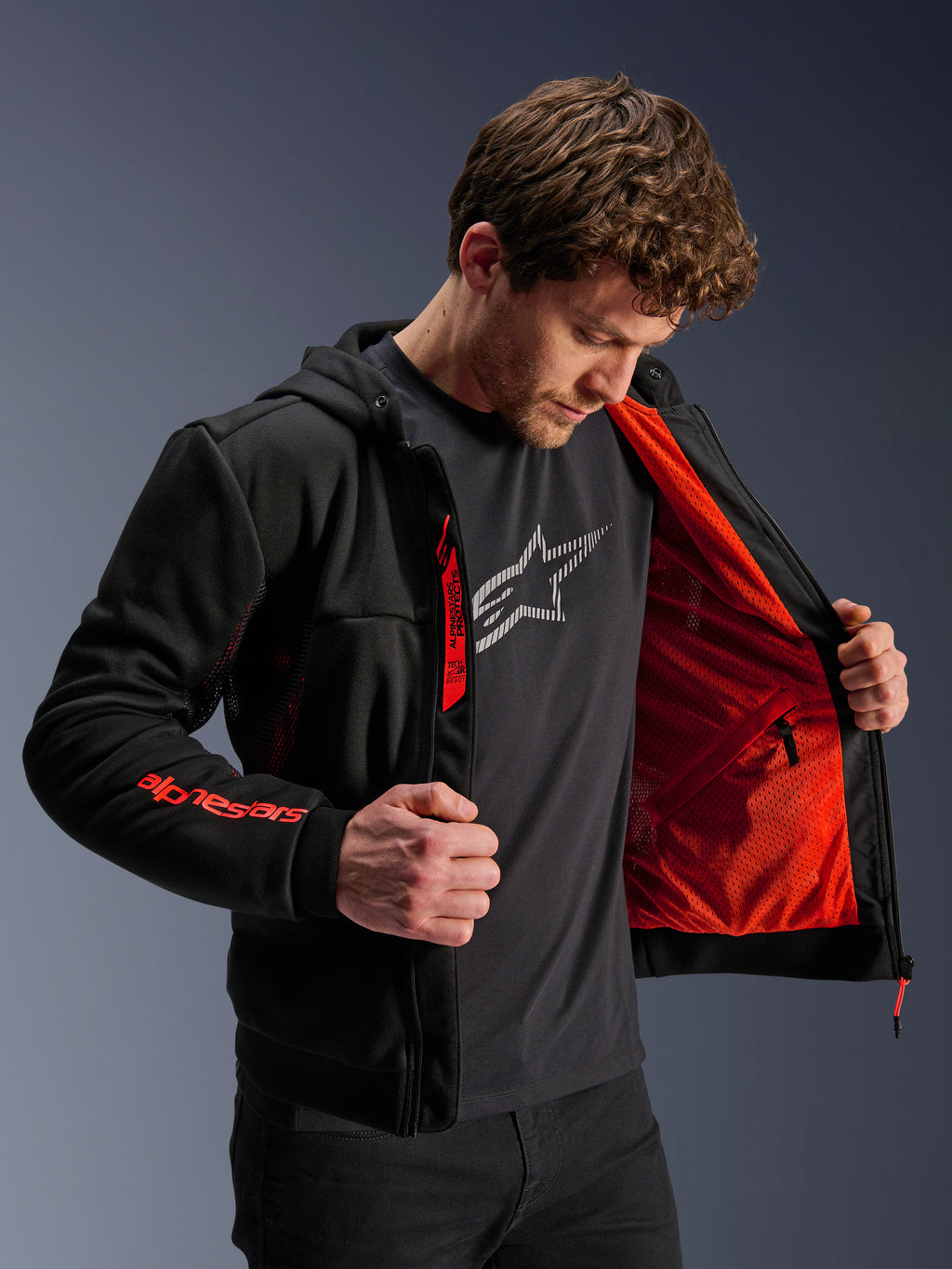 Hoodie Alpinestars Chrome Air Sport em Preto e Vermelho Fluo, modelo masculino a abrir o casaco para mostrar o forro em malha vermelha vibrante, logótipo Alpinestars na manga direita e no fecho do peito, casaco de motociclismo urbano casual concebido para o fluxo de ar.