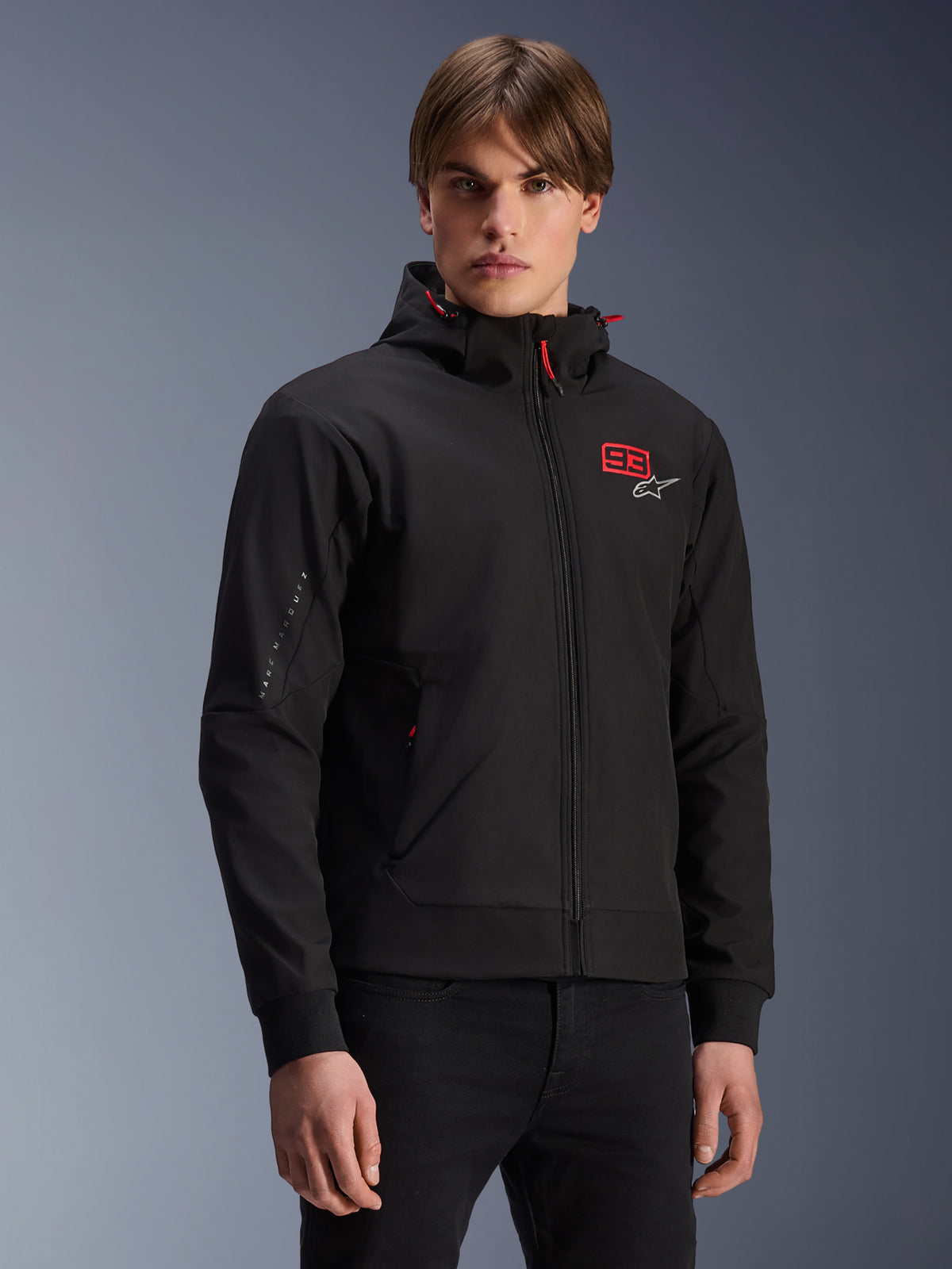 Alpinestars MM93 Radium Tech Hoodie, casaco de motociclismo urbano preto com fecho de correr, logótipo 93 vermelho e Alpinestars branco no peito, modelo com calças pretas, capuz removível com cordões vermelhos e marca na manga, design casual inspirado no desporto motorizado