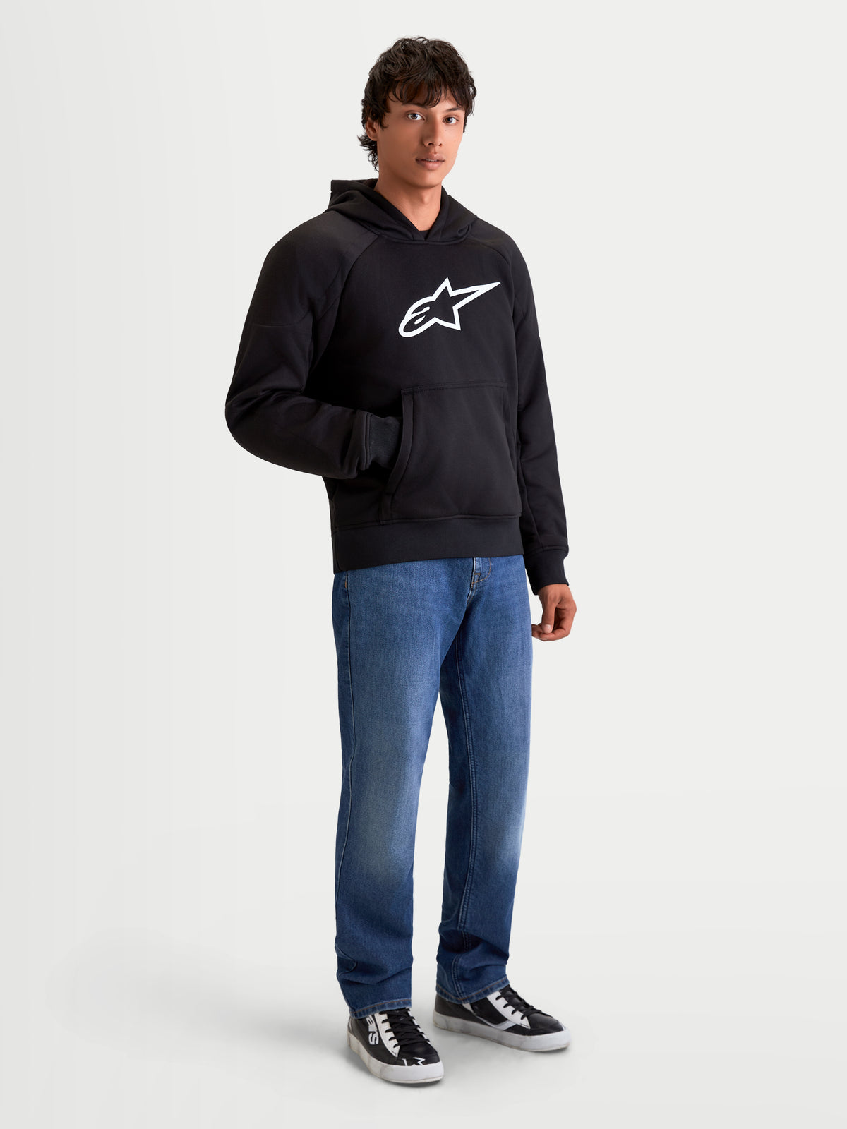 Alpinestars Ageless Hoodie, casaco de moto urbano para homem em preto, vista frontal num modelo com calças de ganga casuais, com um logótipo Astar branco proeminente no peito e reforços internos contra abrasão