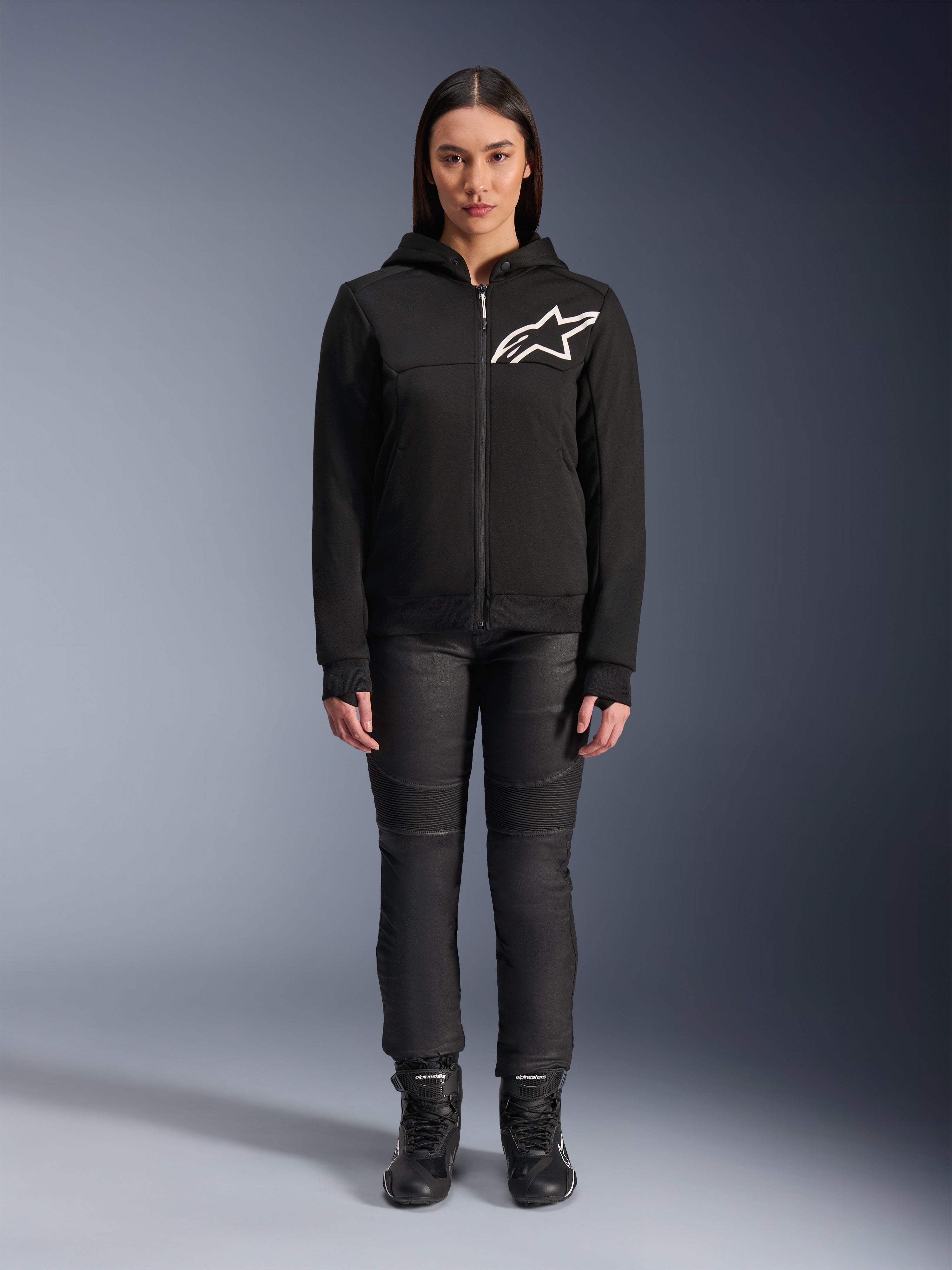Mulher a usar o Alpinestars Stella Chrome V2 Sport Hoodie, preto com logótipo Astars branco no peito, combinado com calças técnicas de condução pretas e botas, design de vestuário urbano para motociclismo