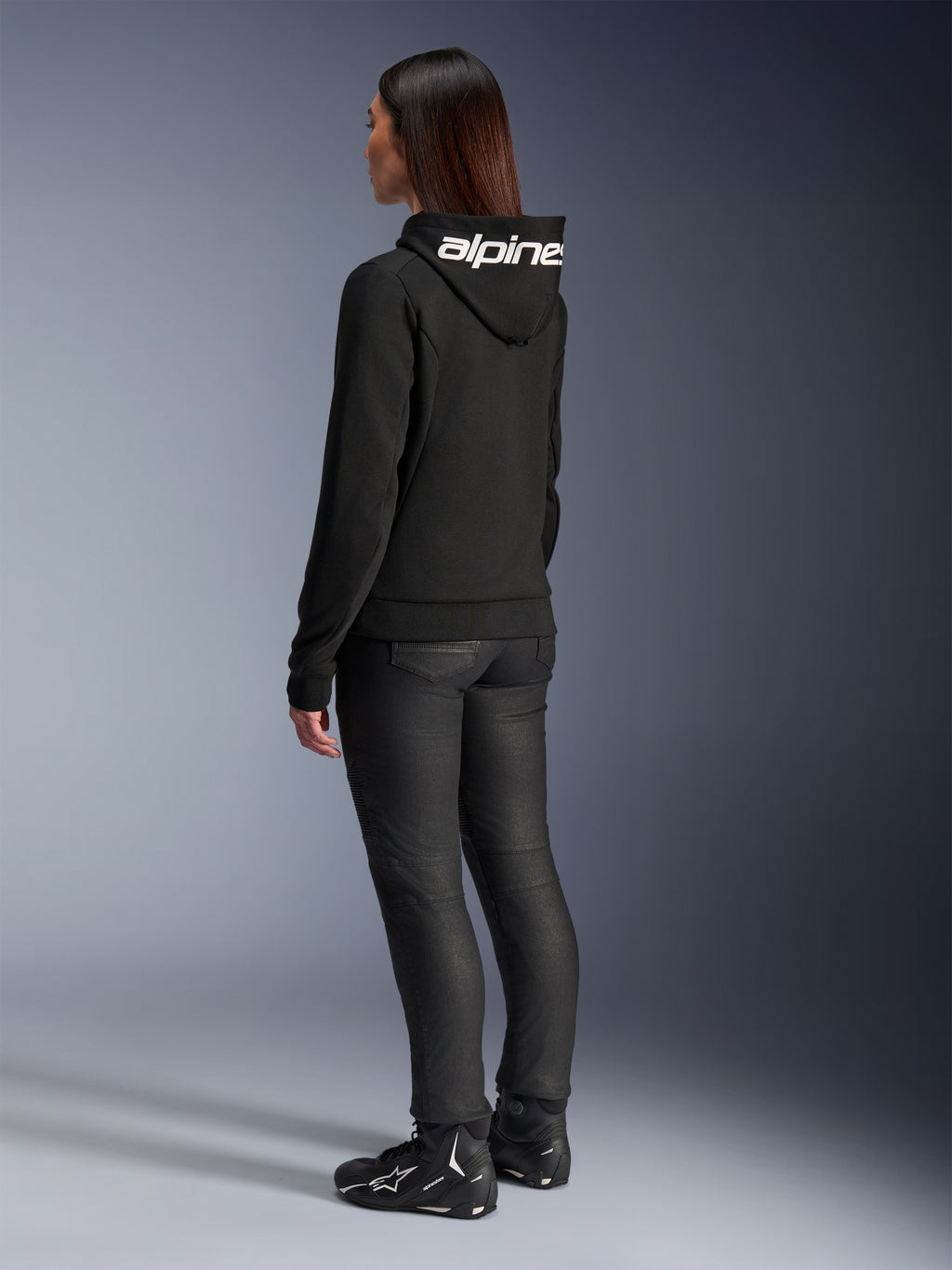 Mulher com o Hoodie Alpinestars Stella Chrome V2 Sport, vista traseira, hoodie de motociclismo preto com logótipo Alpinestars branco no capuz, combinado com calças técnicas de condução pretas e calçado de motociclismo preto, equipamento casual para deslocações urbanas