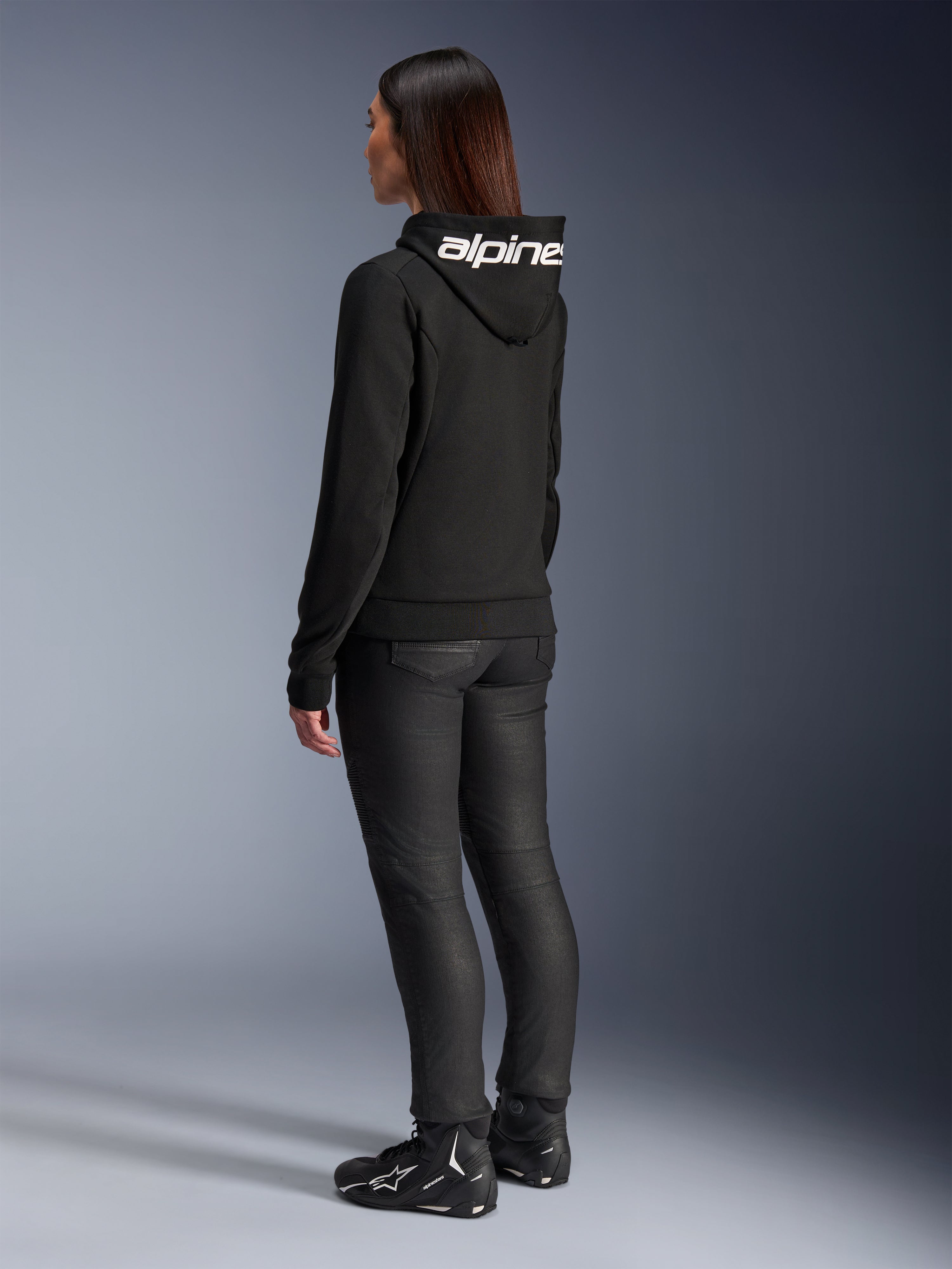 Mulher com o Hoodie Alpinestars Stella Chrome V2 Sport, vista traseira, hoodie de motociclismo preto com logótipo Alpinestars branco no capuz, combinado com calças técnicas de condução pretas e calçado de motociclismo preto, equipamento casual para deslocações urbanas