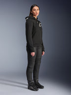 Mulher a usar Hoodie Alpinestars Stella Chrome V2 Sport em preto, com logótipo Alpinestars em branco, combinado com calças e botas de motociclismo pretas, equipamento de condução urbana para mulher com um ajuste atlético casual