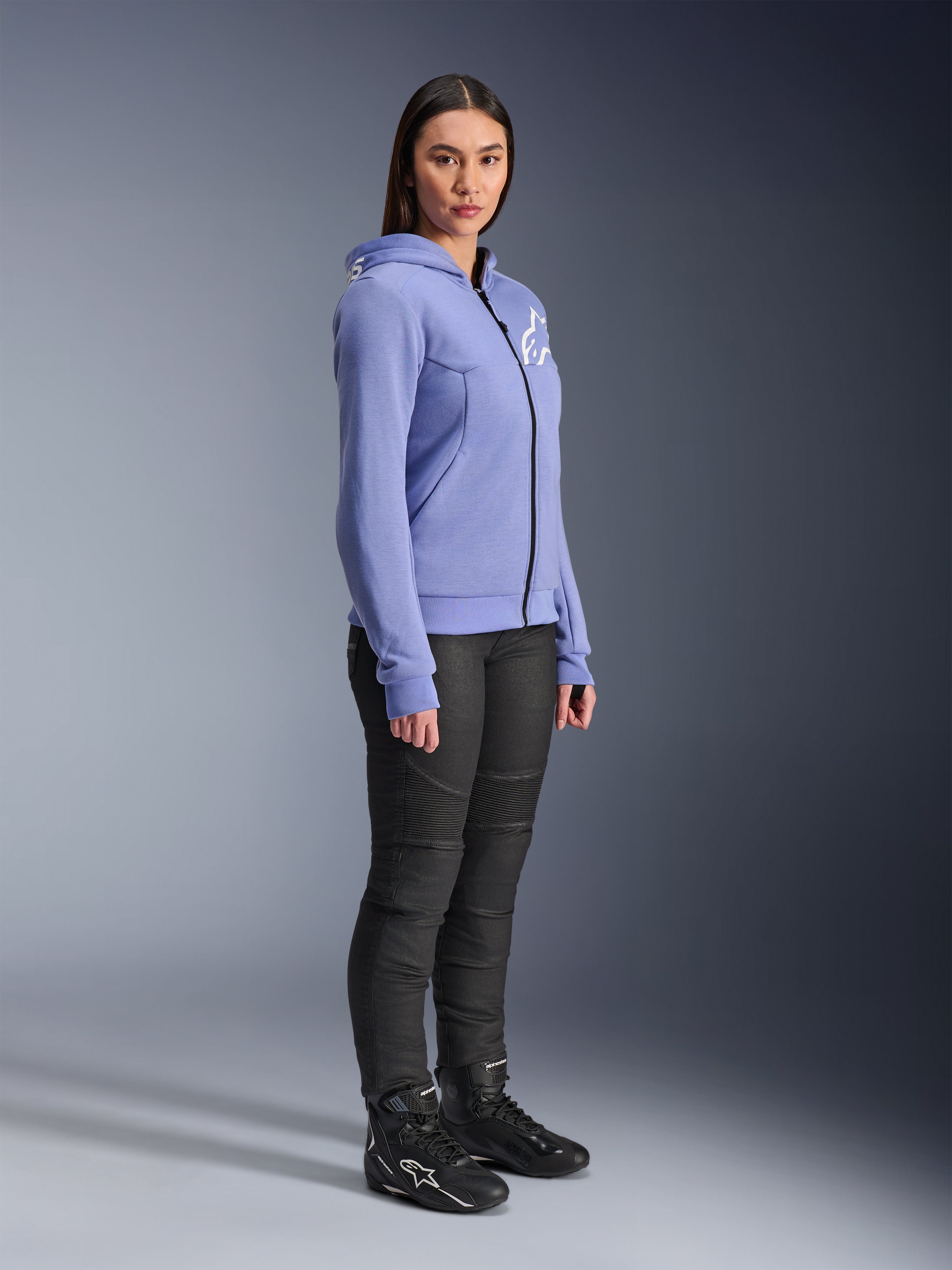 Alpinestars Hoodie Feminino Stella Chrome V2 Sport, hoodie de motociclismo em azul-violeta com logótipo Alpinestars branco no peito, vestido por uma modelo com calças de condução pretas e botas de motociclismo pretas, vestuário de condução urbana para mulher