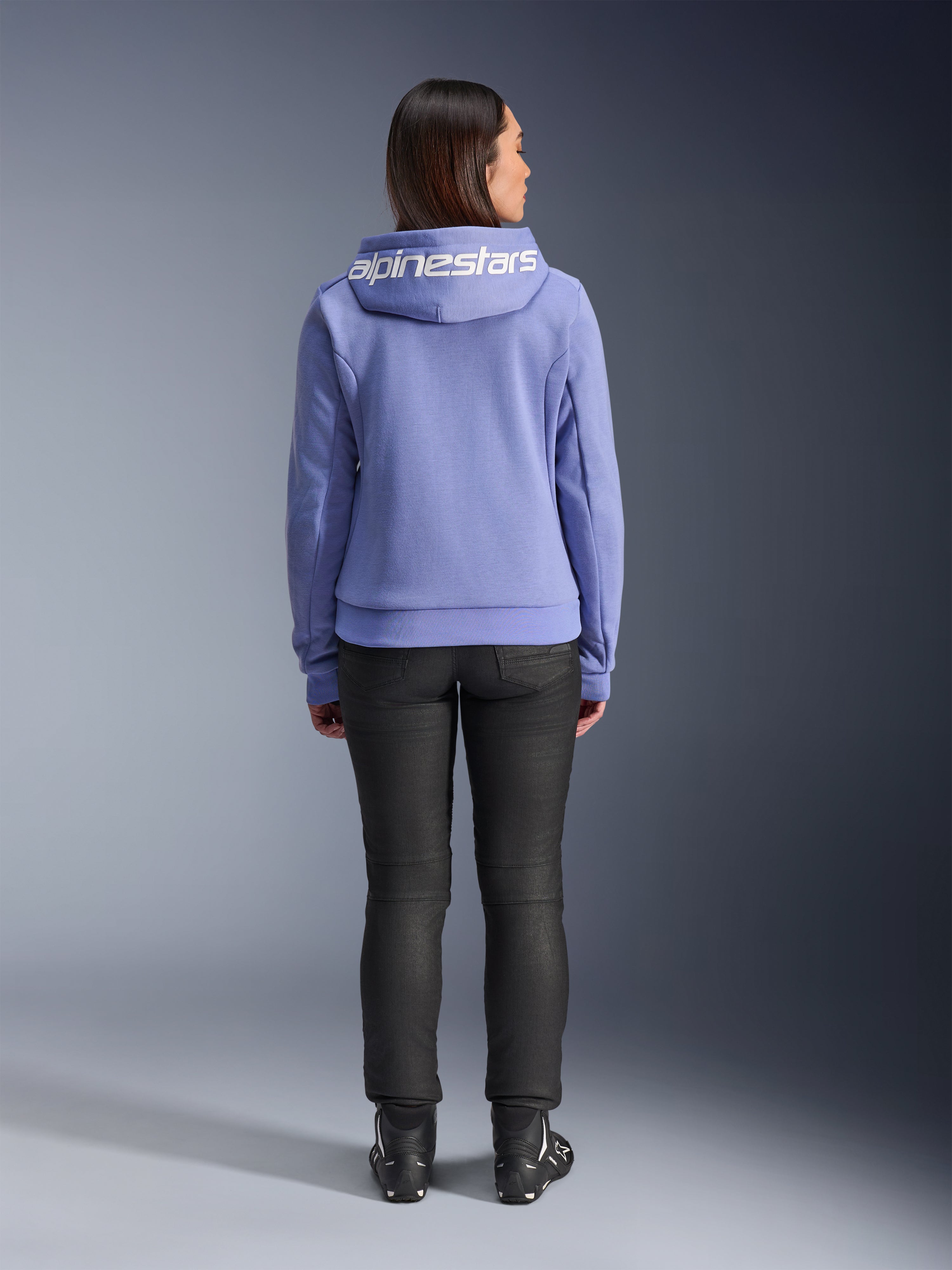 Alpinestars Stella Chrome V2 Sport Hoodie, azul violeta, vista traseira de uma mulher a usar o hoodie com o logótipo Alpinestars branco no capuz, calças pretas e botas de motociclismo, vestuário urbano casual para motociclismo