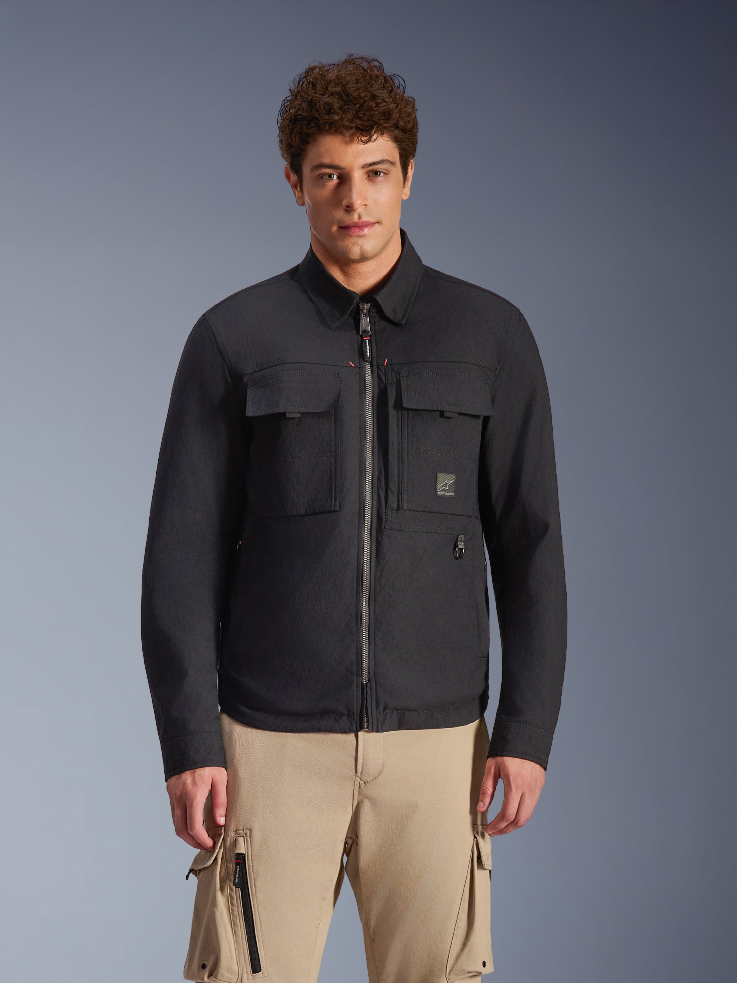 Alpinestars Bryon Water Resistant Overshirt, preto, casaco de motociclismo urbano com estilo inspirado em workwear, com dois bolsos cargo grandes no peito e fecho de correr frontal integral, em lona encerada, usado por um modelo com calças cargo bege para um visual utilitário contemporâneo