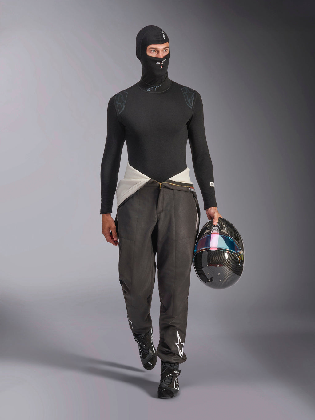 Balaclava Alpinestars ZX Evo V3, preta, usada por um modelo num conjunto completo de roupa interior técnica de competição, incluindo uma camisola de manga comprida a condizer e calças de competição cinzentas, segurando um capacete de fibra de carbono com viseira iridescente, equipamento profissional de automobilismo