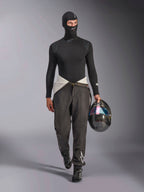 Balaclava Alpinestars ZX Evo V3, preta, usada por um modelo num conjunto completo de roupa interior técnica de competição, incluindo uma camisola de manga comprida a condizer e calças de competição cinzentas, segurando um capacete de fibra de carbono com viseira iridescente, equipamento profissional de automobilismo